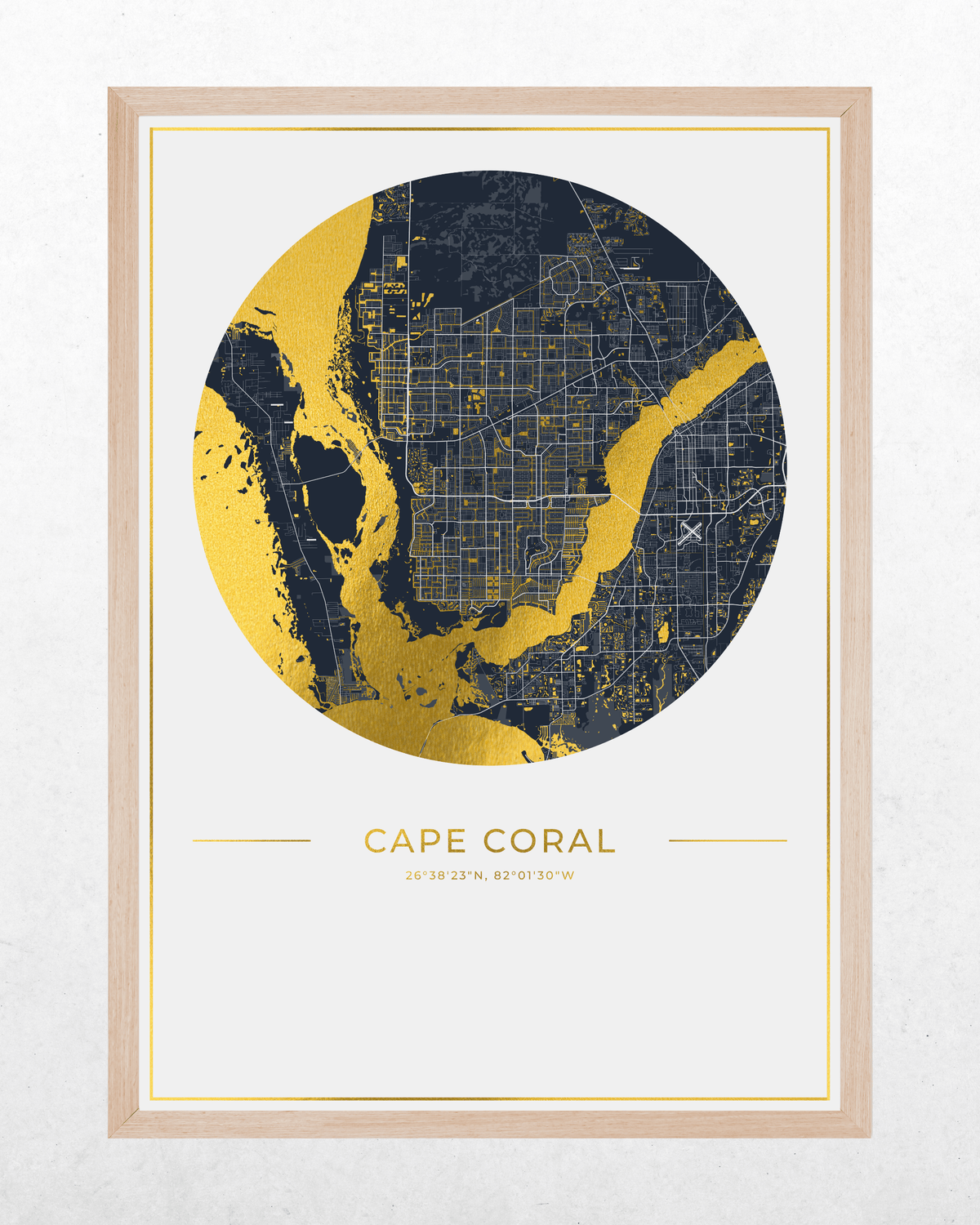 A framed circular map print of Cape Coral, Florida, featuring a navy blue and gold map design with coordinates printed below, set in a light wood frame against a textured white wall.