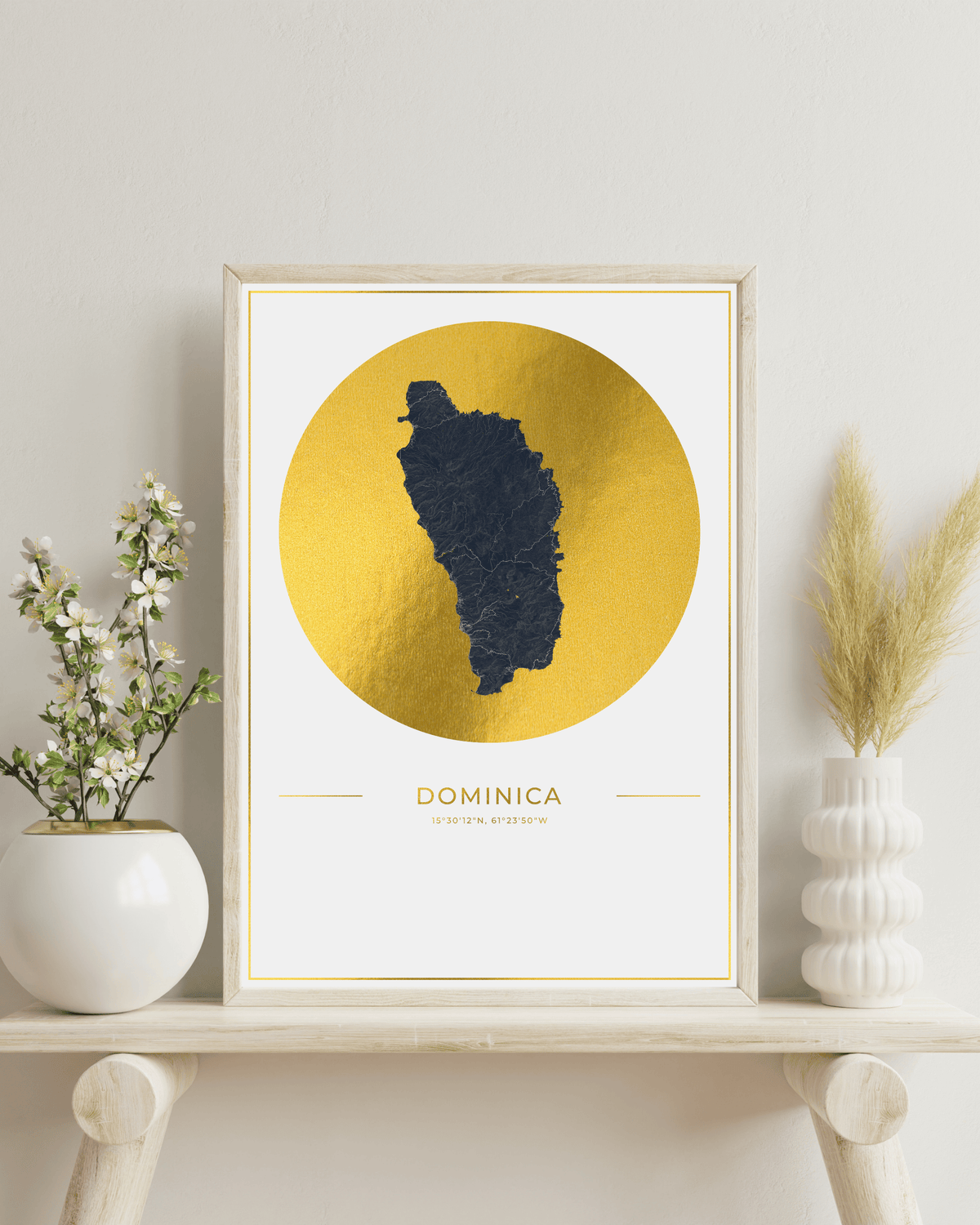A framed art print featuring a dark navy silhouette map of Dominica on a textured gold circular background, styled on a light wood shelf with white vases and dried grass.