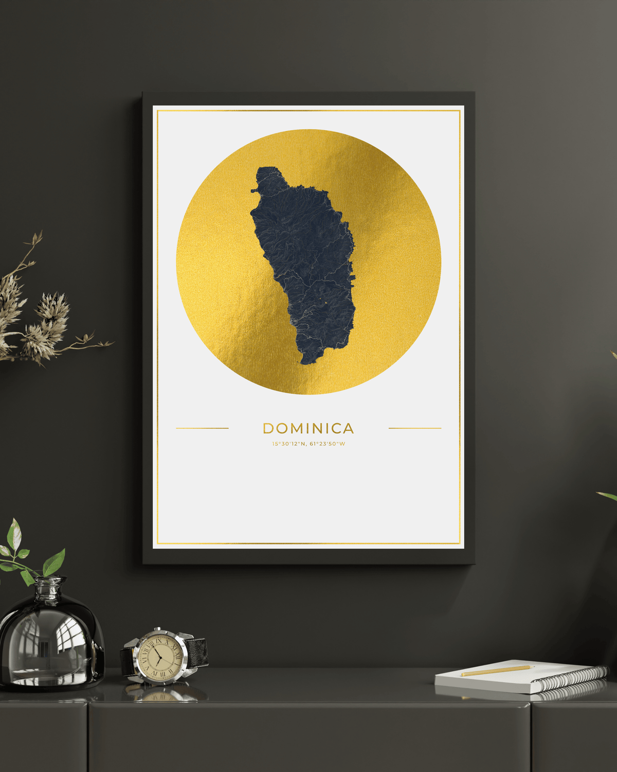 A framed wall art print featuring a dark navy silhouette map of Dominica over a large textured gold circle on an off-white background, displayed on a dark gray wall above a console table with decorative objects.