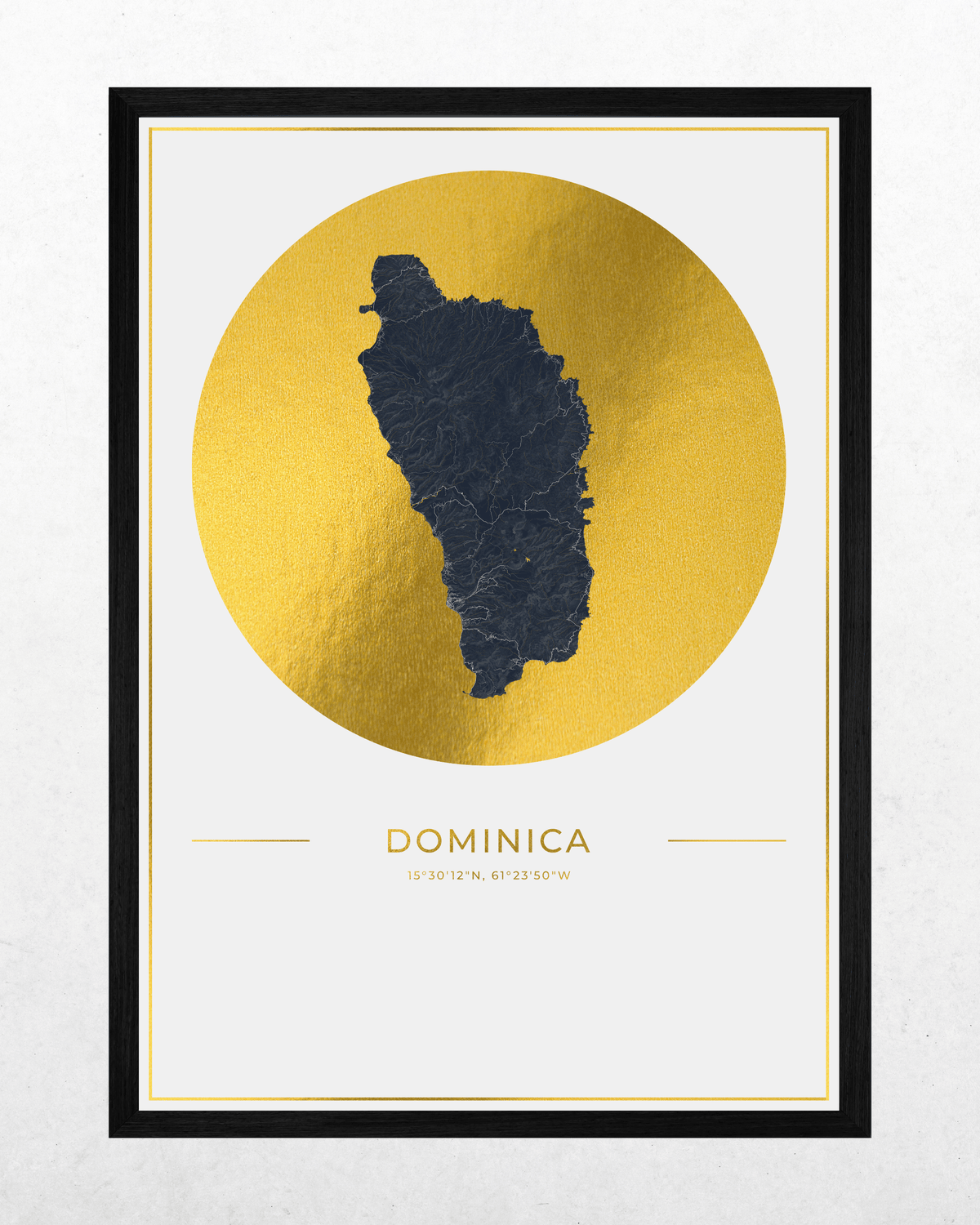 A framed art print featuring a dark charcoal map silhouette of the island of Dominica centered inside a large textured gold circle, with the word 'DOMINICA' and coordinates printed below on a white background. The print is enclosed in a thin white mat and a black frame, shown against a light gray wall.