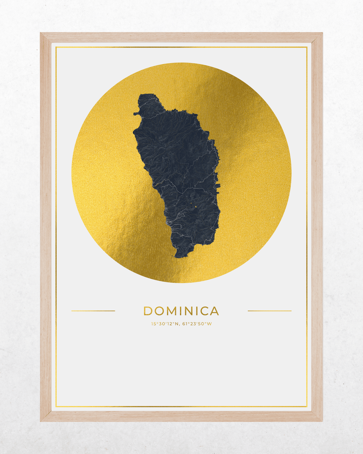 A framed art print featuring a dark charcoal map silhouette of the island of Dominica centered inside a large textured gold circle, with the word 'DOMINICA' and coordinates printed below on a white background. The print is enclosed in a natural wood frame, shown against a light gray wall.