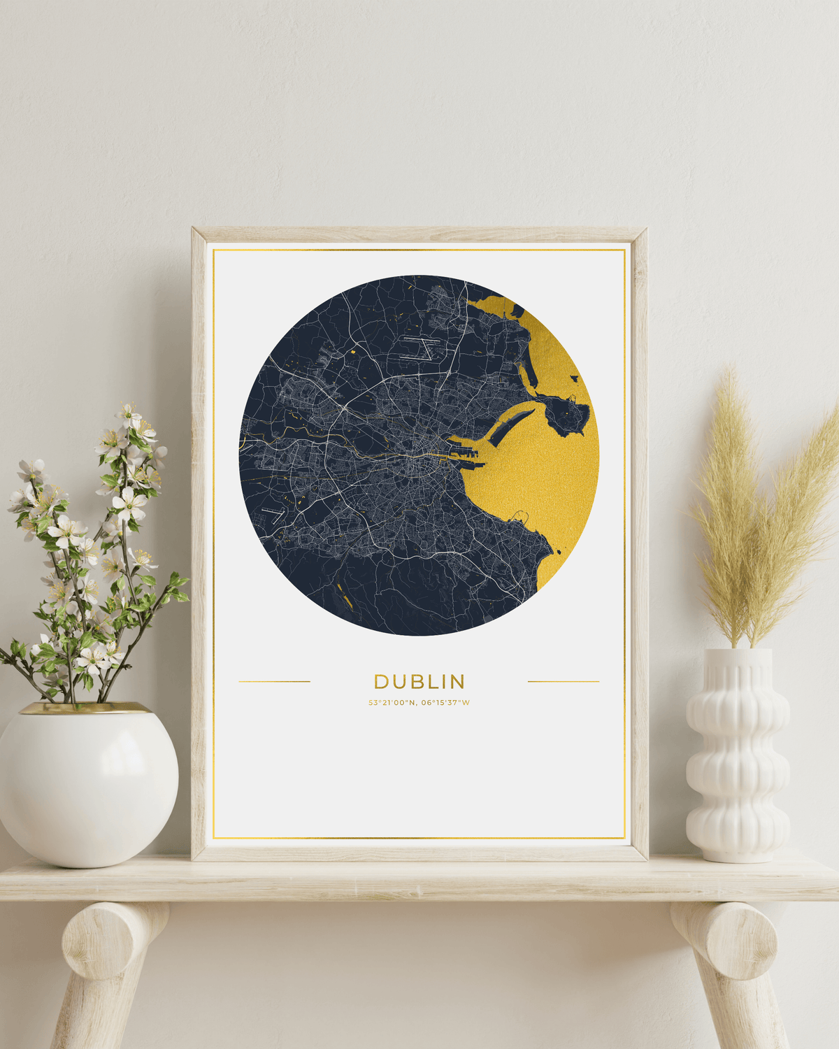 A framed circular city map print of Dublin, Ireland, styled on a light wood shelf with white ceramic vases and dried grass stems against a neutral wall.