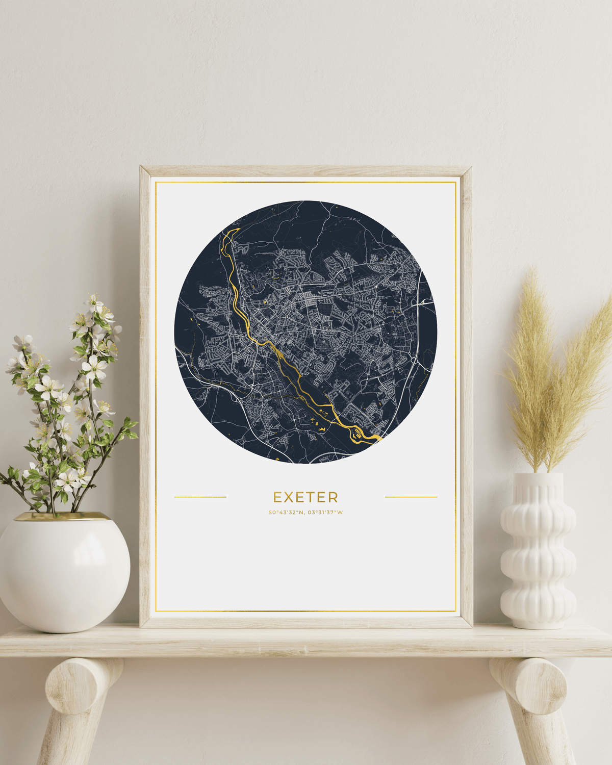 A framed circular city map print of Exeter, Devon, England, styled on a light wood shelf with white ceramic vases containing greenery against a neutral wall.