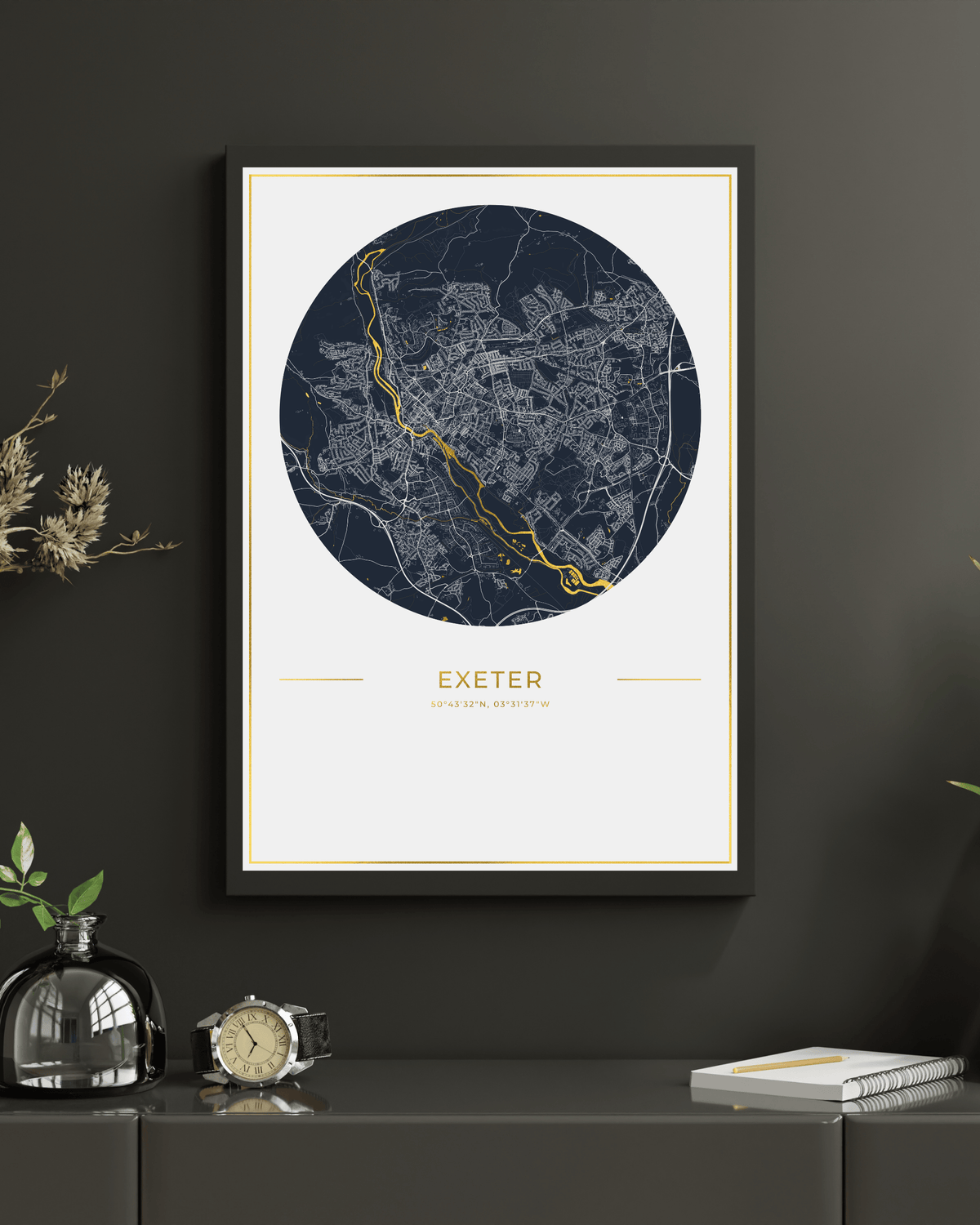 A framed art print of a circular street map of Exeter, England, featuring a navy blue map with white street lines and a highlighted gold route. The print is displayed on a dark gray wall above a dark gray console table with decorative items including a glass bottle, a clock, and a notebook.
