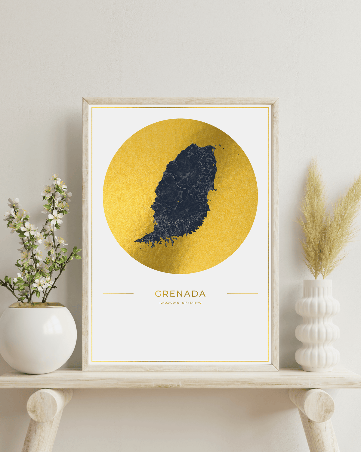 A framed art print featuring a dark blue map silhouette of Grenada on a textured gold circular background, styled on a light wood shelf with white vases and dried grass.