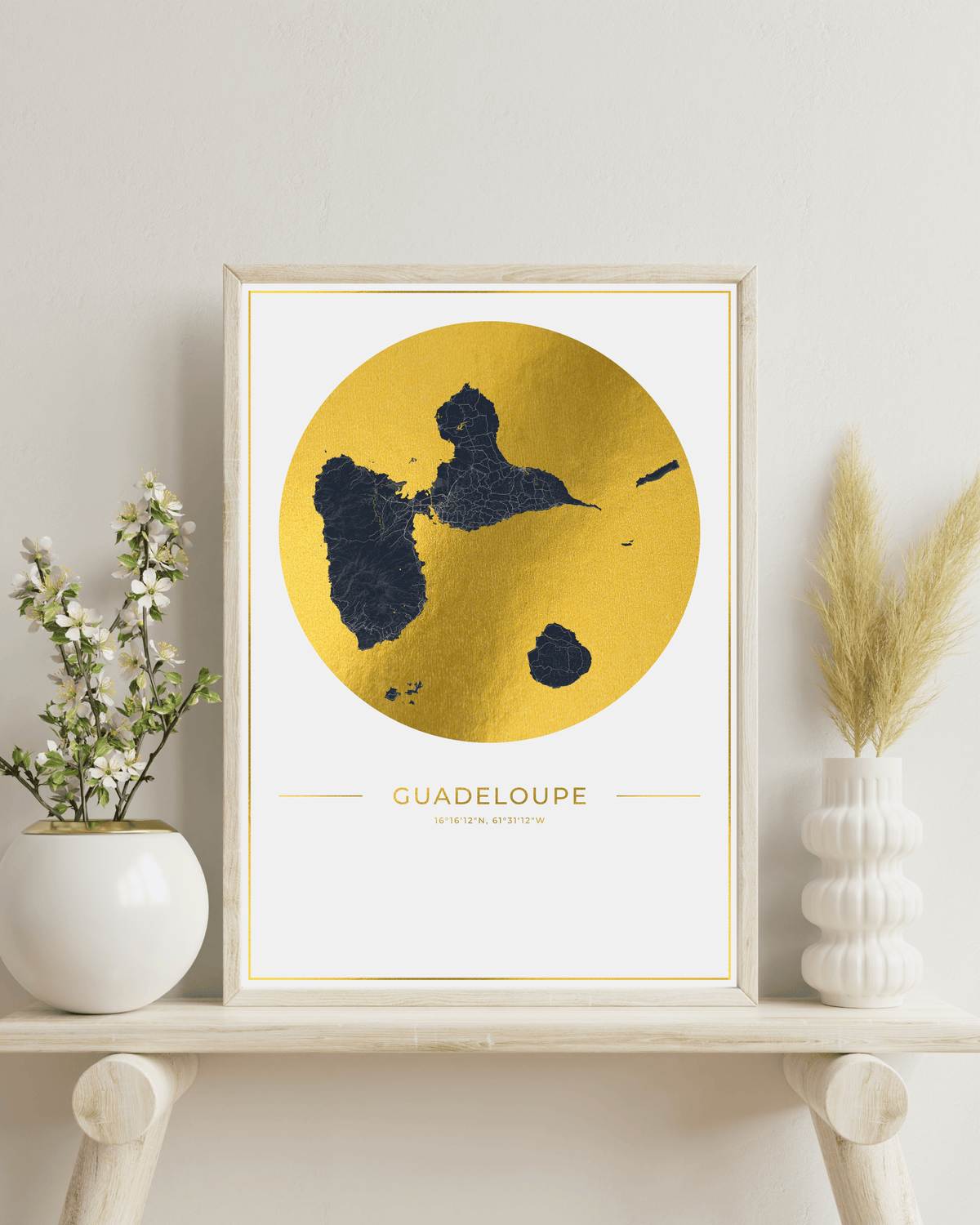 Framed wall art print featuring a gold map of Guadeloupe with dark navy landmasses, displayed on a light wood shelf between white ceramic vases against a neutral wall.