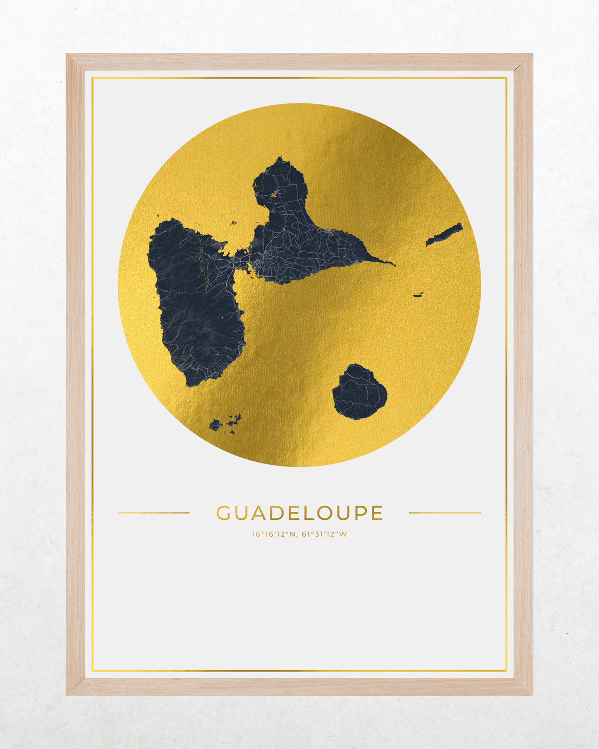 A framed art print of Guadeloupe featuring a gold foil map of the island with dark blue details, set in a light wood frame against a textured white wall.