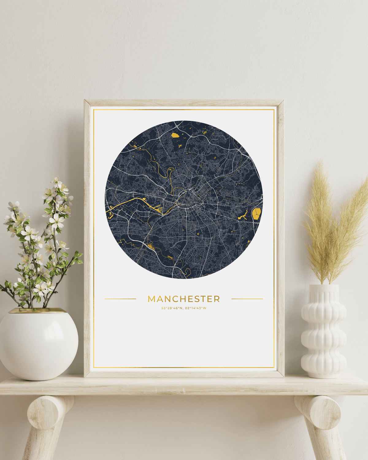 A framed circular city map print of Manchester, UK, styled on a light wood shelf with white ceramic vases and dried pampas grass against a neutral wall.