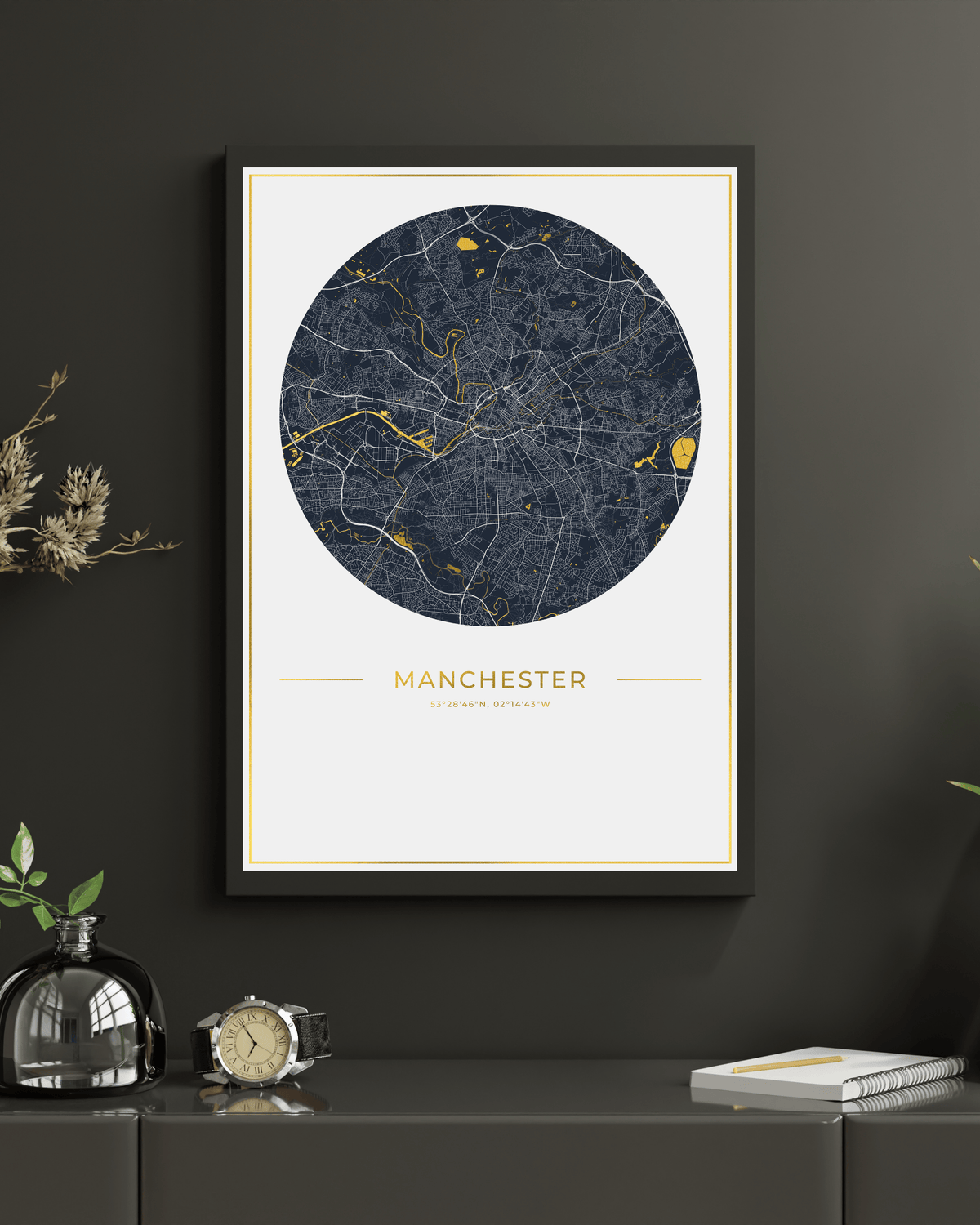 A framed circular street map print of Manchester, UK, featuring a dark navy blue map with gold highlights on an off-white background, displayed on a dark gray wall above a console table with decorative items.