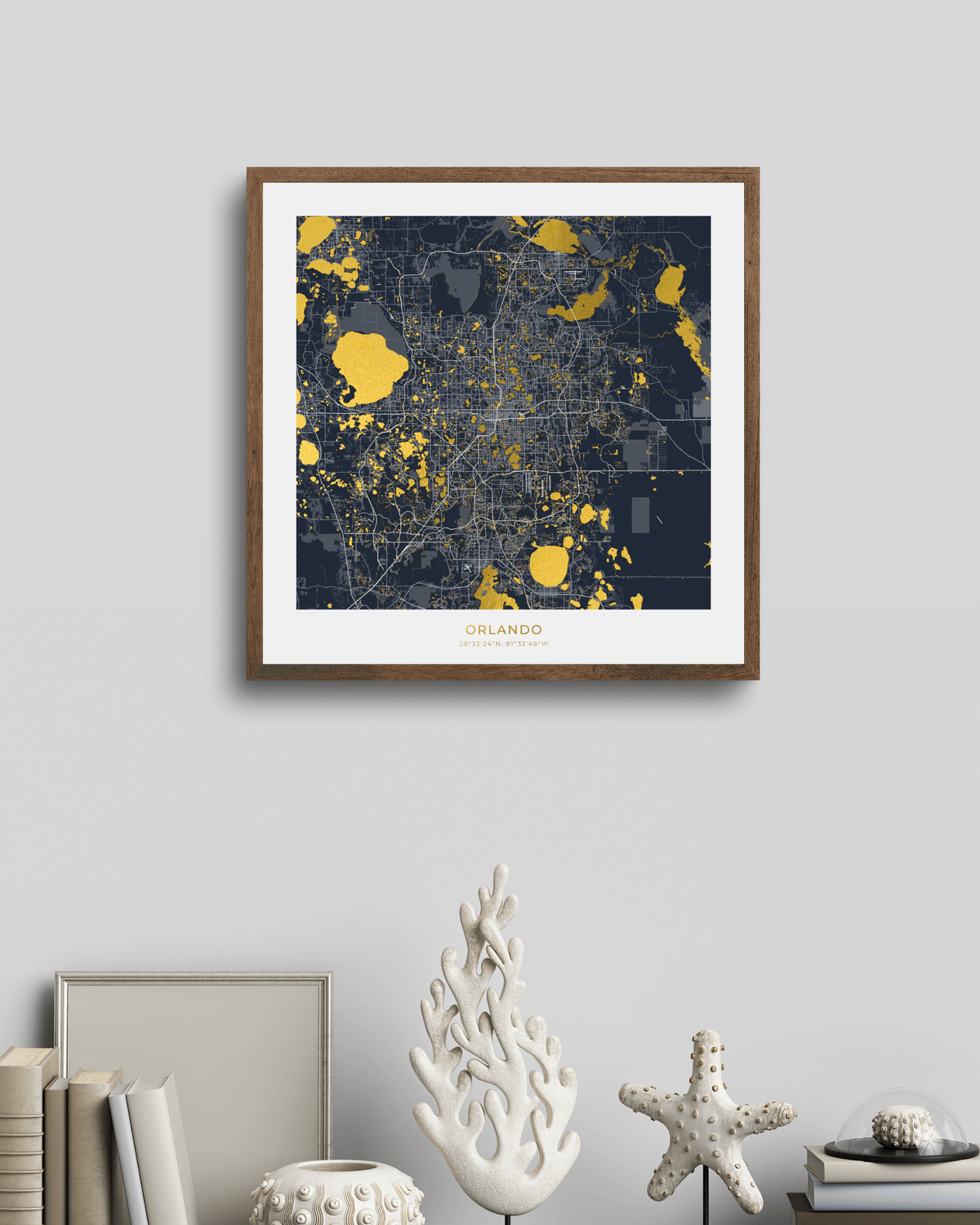 A framed city map print of Orlando, Florida, featuring a dark navy blue background with gold highlights and a white mat board, housed in a brown wood frame. The frame is displayed on a light gray wall above a styled shelf with books and decorative coral figurines.