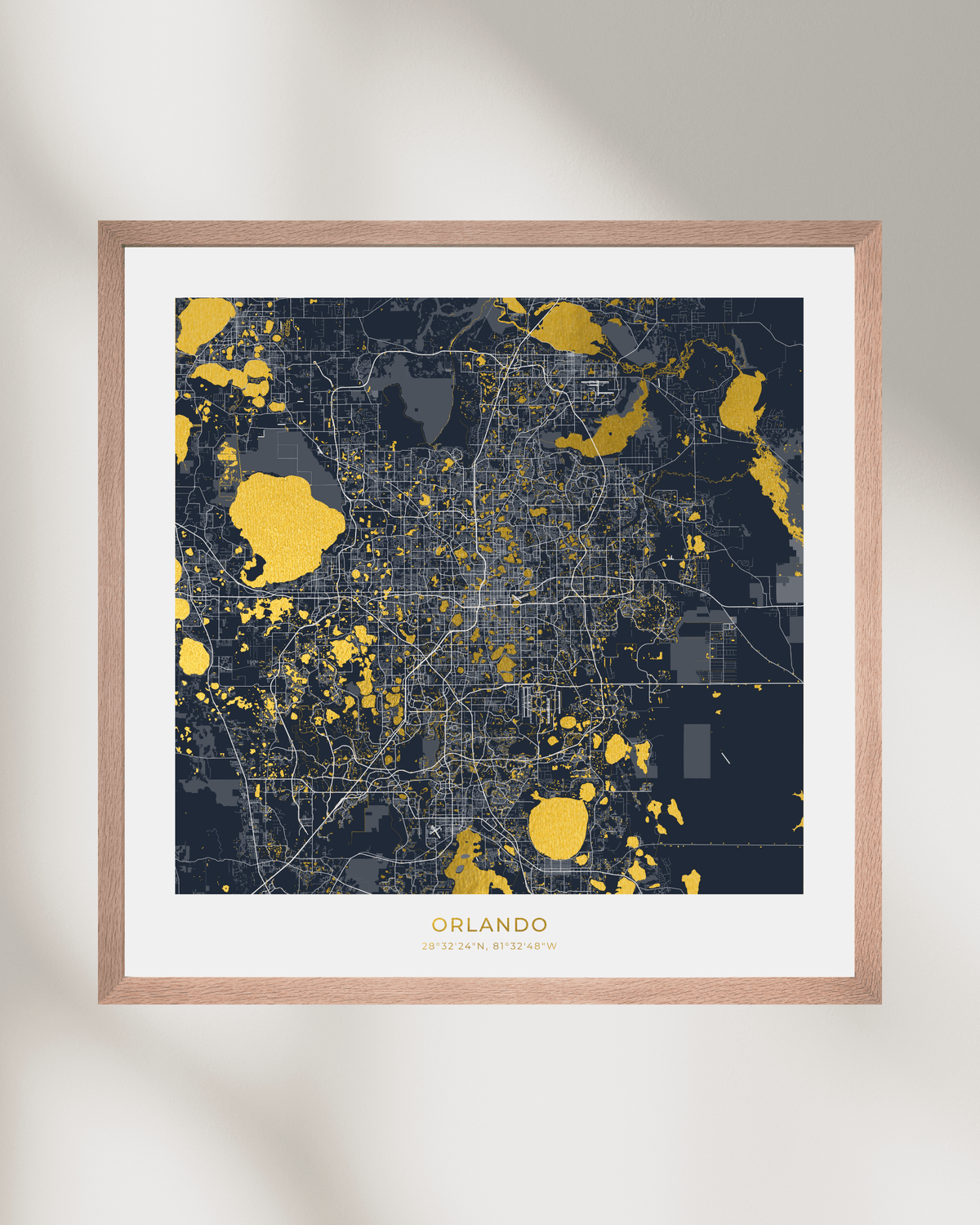 A framed city map print of Orlando, Florida, featuring a dark navy blue and gray map with gold highlights, set in a light wood frame with a white mat against a neutral wall.