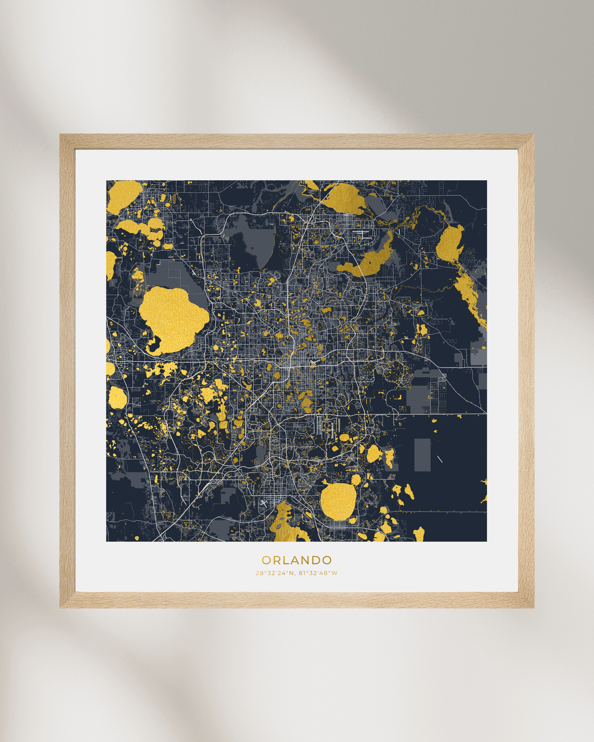 A framed city map print of Orlando, Florida, featuring a dark navy and gray street layout highlighted with gold highlights, set in a light natural wood frame with a white mat on a neutral wall.