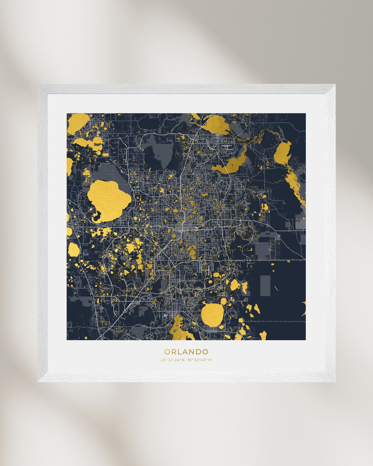 A framed city map print of Orlando, Florida, featuring a dark navy and gray street grid with gold highlighted areas of water, presented in a white frame against a light beige wall with soft shadows.