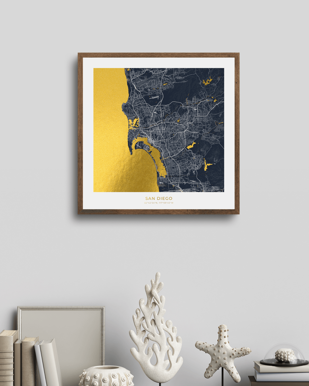 A framed San Diego city map wall art print hangs on a light gray wall above a styled shelf with books, a white coral sculpture, a starfish, and small decorative objects. The print features a navy street map with a large gold coastal area and white matting, in a brown wood frame.
