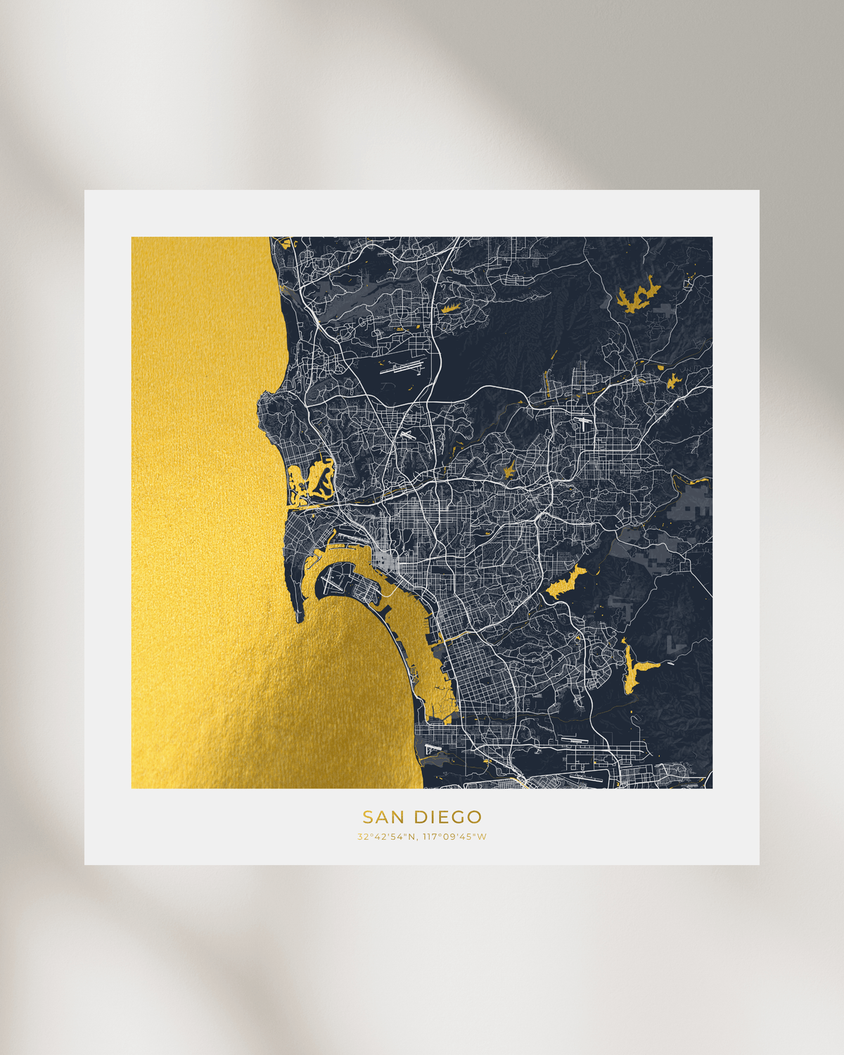 A square city map print of San Diego, California, featuring a dark navy street grid and coastline with metallic gold highlights on the water and text, displayed on a white background with soft shadows.