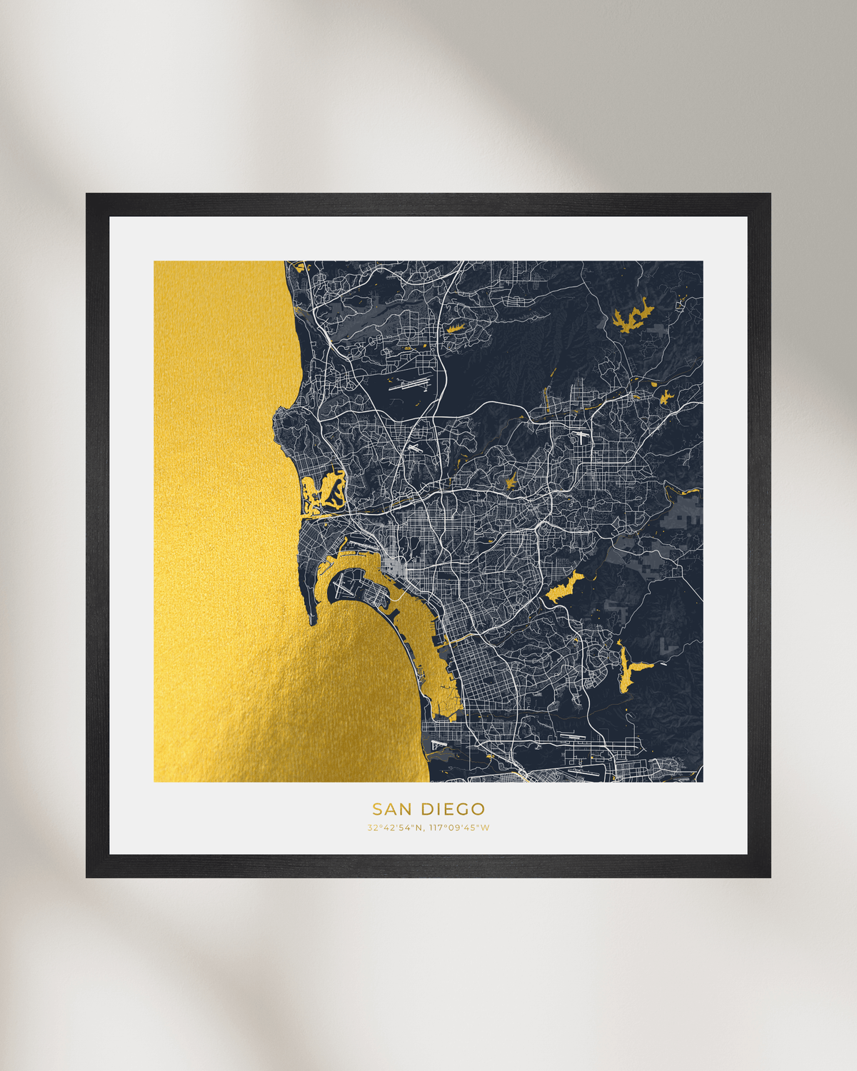 Framed San Diego city map wall art with a black frame, white mat, and a gold-and-dark map design, shown hanging on a light wall.