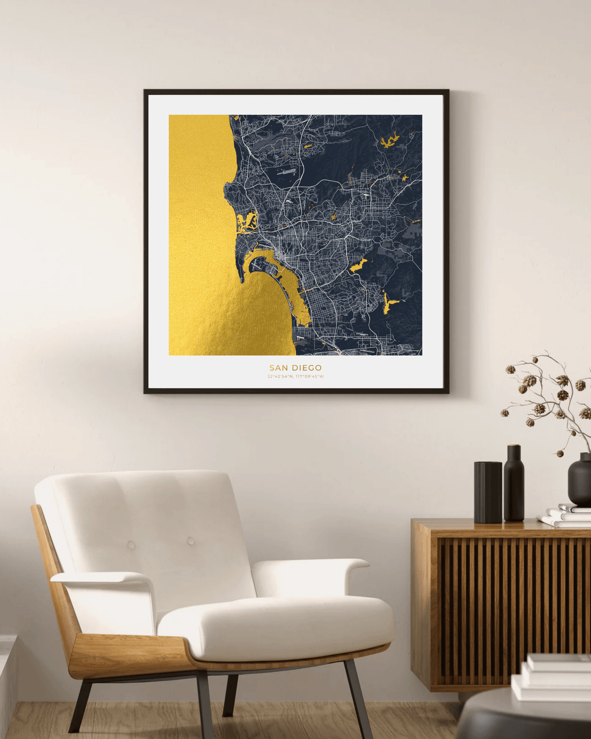A framed San Diego city map print with a gold foil texture on the water and dark navy land, displayed on a light beige wall above a modern white armchair and a wooden sideboard.