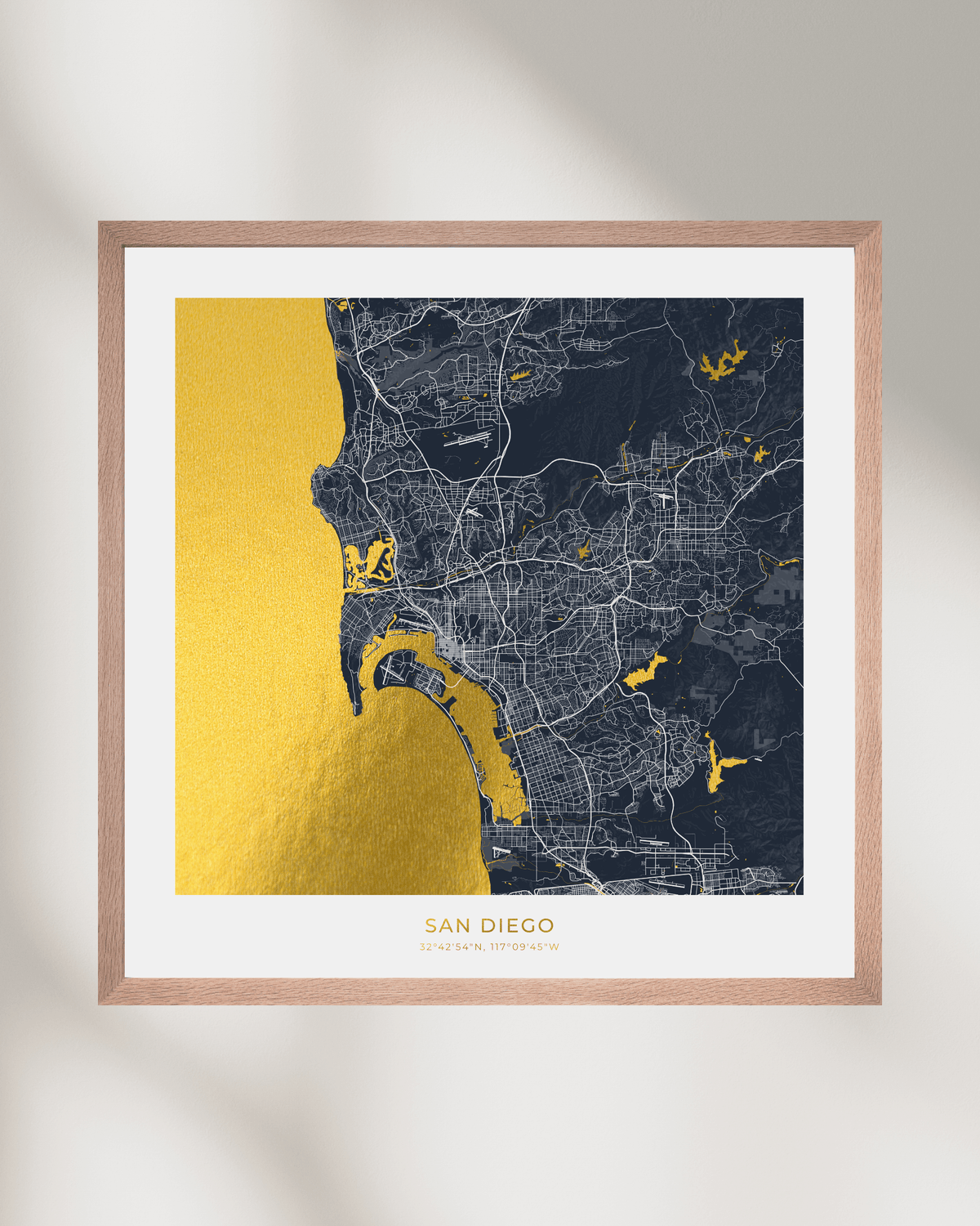 Framed San Diego city map wall art with a gold foil coastline and highlights, dark navy street map background, white mat, and a light wood frame on a softly lit neutral wall.