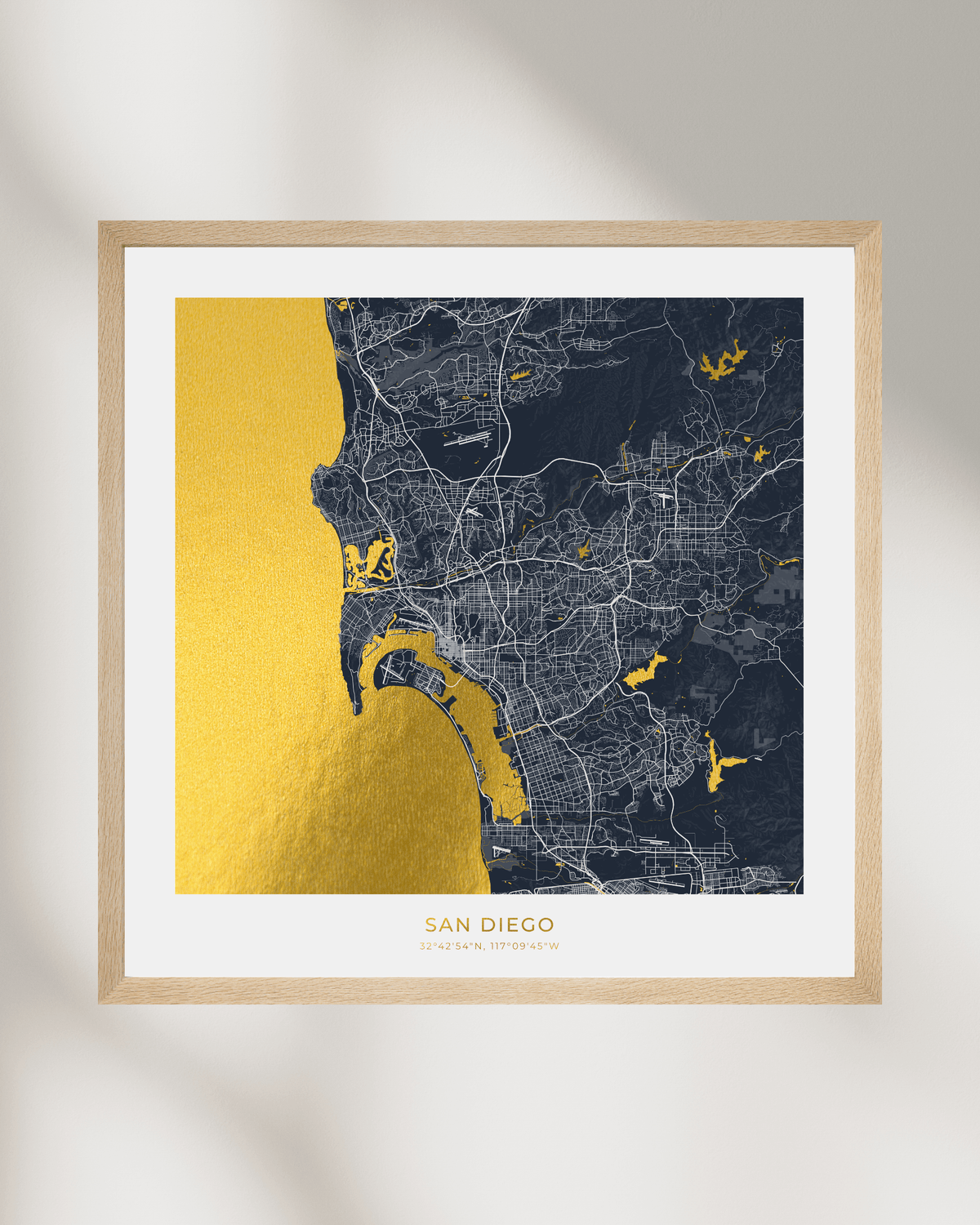 A framed San Diego city map art print with a gold foil coastline and water features, a dark navy street map, and a light wood frame, displayed against a neutral wall with soft shadows.