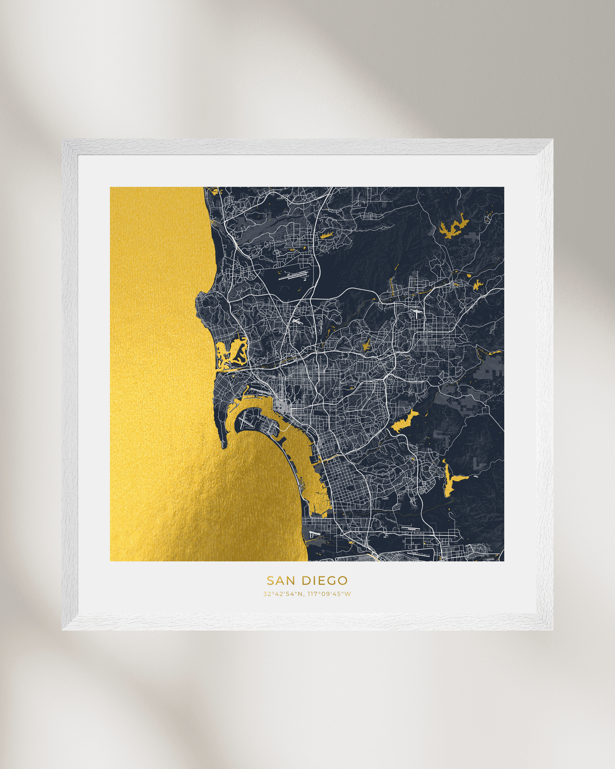 A framed San Diego city map art print with a textured gold foil coastline and water features, set against a dark navy street map background. The print is housed in a clean white frame and displayed on a light gray wall with soft, diagonal shadows.