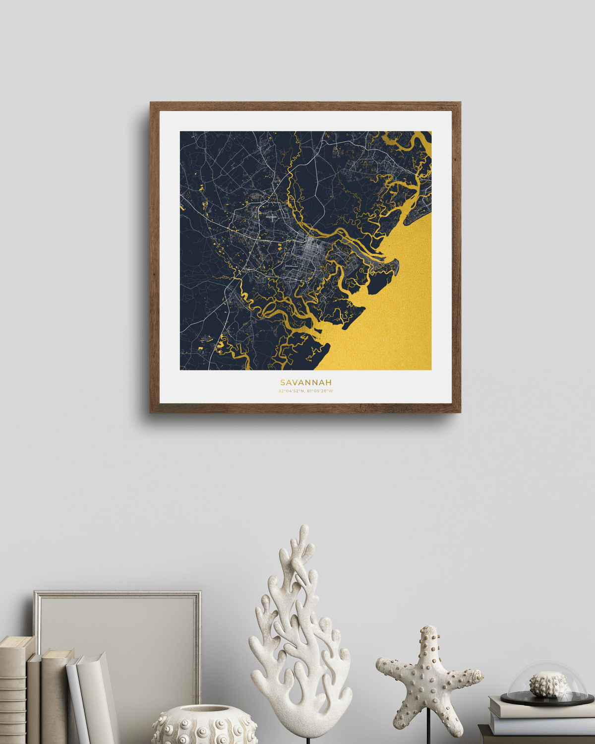A framed map print of Savannah, Georgia, featuring a dark navy blue and gold coastal map design with a white mat and a brown wood frame, displayed on a light gray wall above a styled shelf with books and decorative objects.