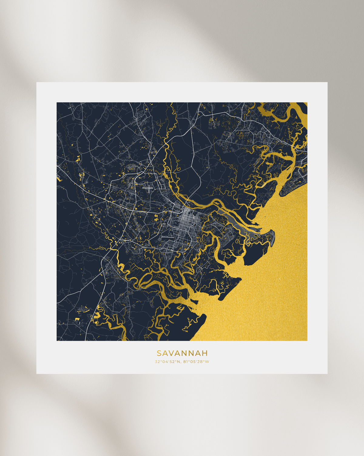 A minimalist city map art print of Savannah, featuring a dark navy blue land mass with white street lines and a textured gold coastline and bay area, displayed on a white paper sheet against a light gray background with soft shadows.