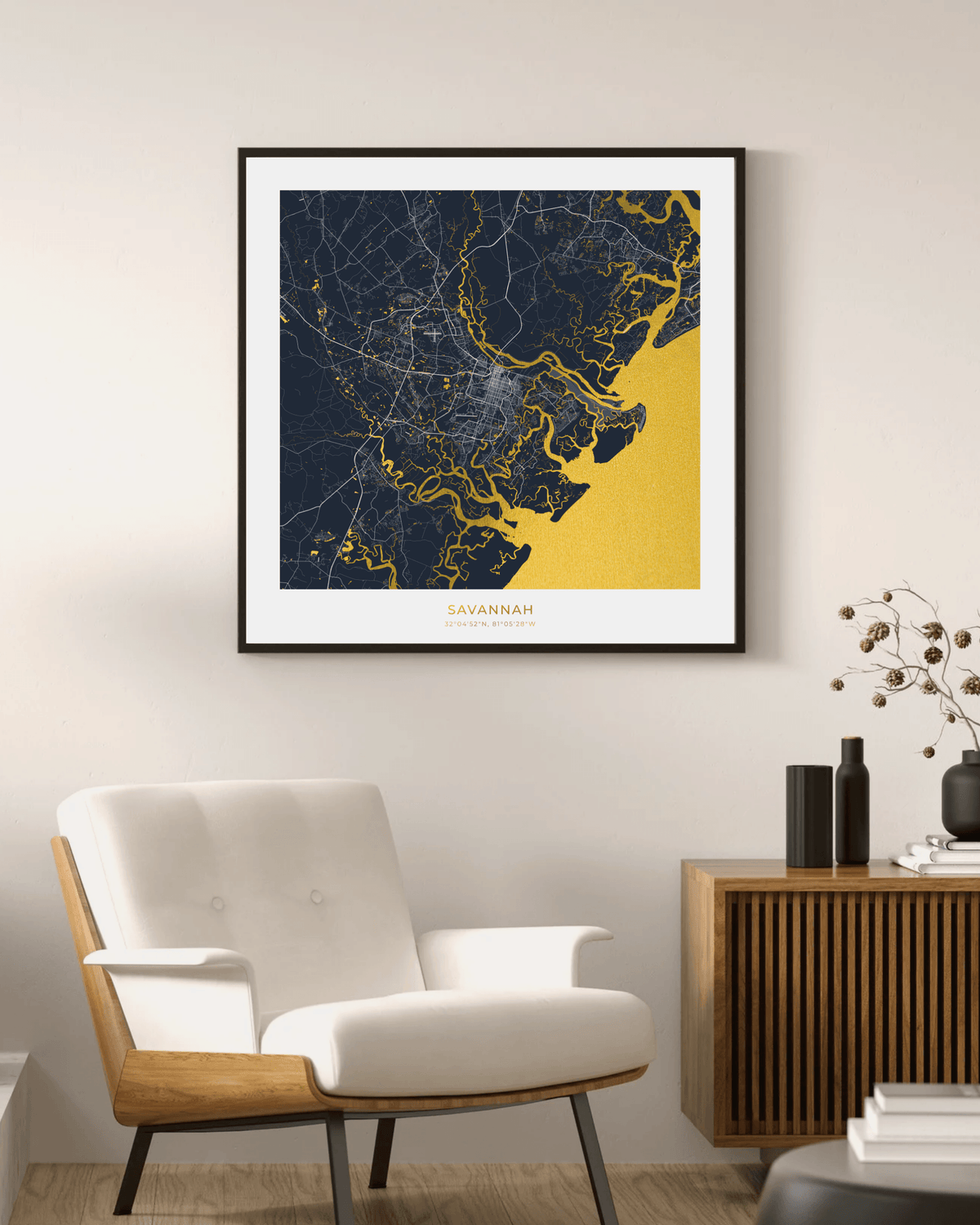 A framed square map print of Savannah, Georgia, featuring a navy blue and yellow coastal design, displayed on a beige wall in a modern living room setting above a white armchair.