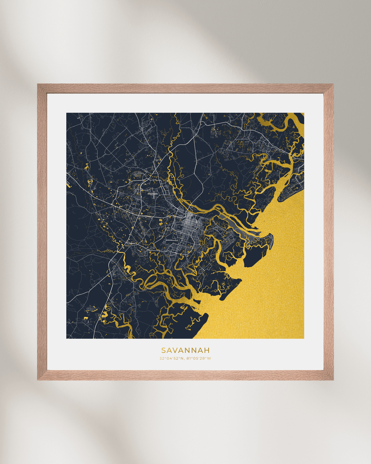 A framed map art print of Savannah, Georgia, featuring a dark navy blue land mass with waterways coloured in gold water, set in a light wood frame with a white mat on a neutral background.