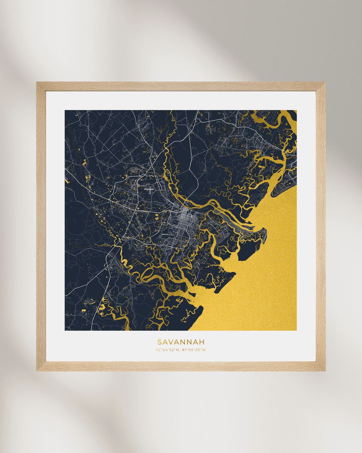 A framed map art print of Savannah, Georgia, featuring a dark navy blue land mass with gold street lines and a large gold body of water, set in a light wood frame against a white wall with soft shadows.