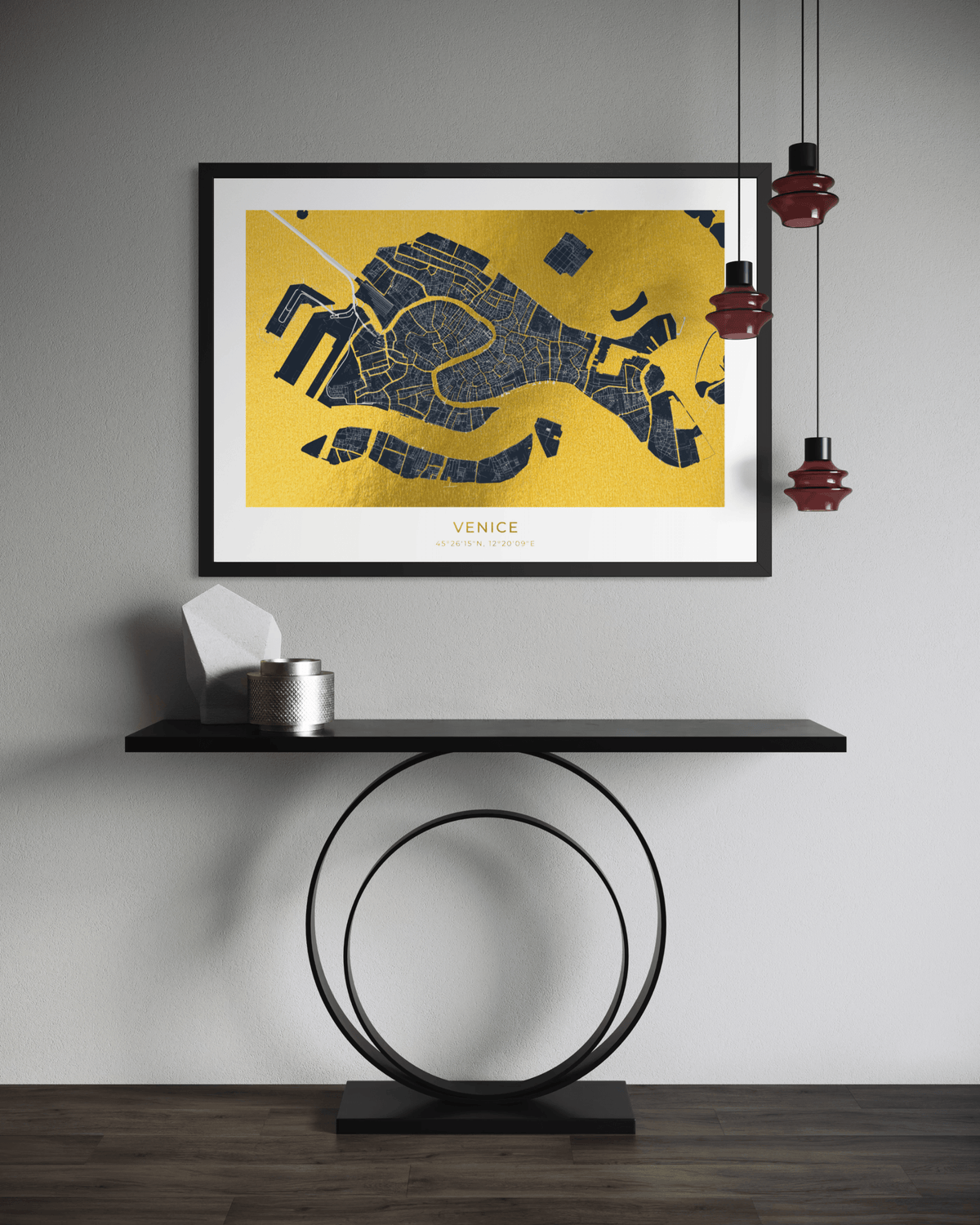 A framed Venice street map art print with a gold background and dark blue map lines, hanging on a light gray wall above a black console table with decorative objects and hanging red pendant lights.