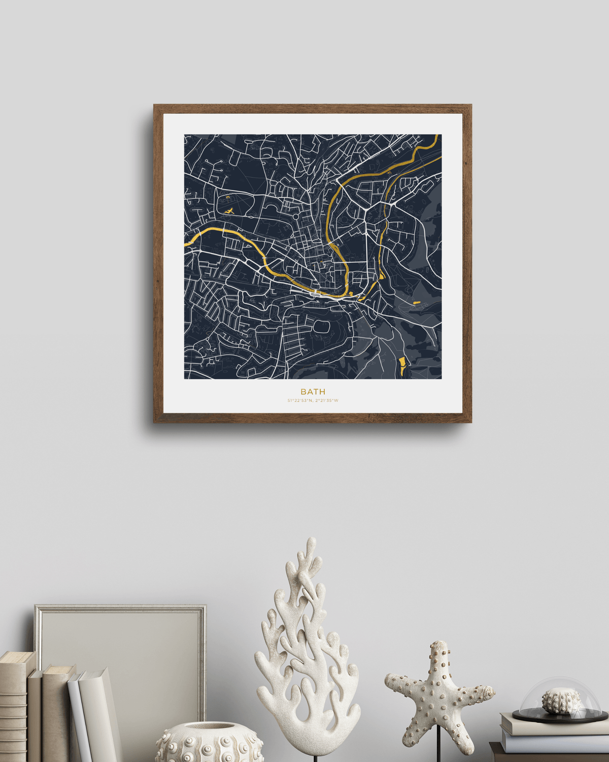 A framed street map art print of Bath, England, featuring a dark navy blue background with white street lines and waterways highlighted in gold foil . The print is housed in a medium brown wood frame with a white mat, hanging on a light gray wall above a styled shelf with books and decorative coral figurines.