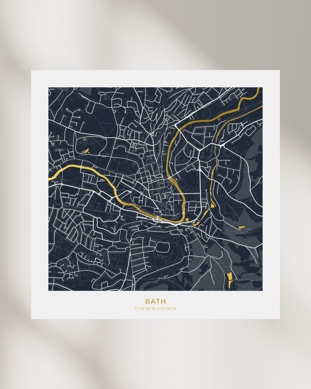 A minimalist city map print of Bath, UK, featuring a dark navy blue background with white street lines and waterways highlighted in gold foil. The print is displayed on a clean white background with soft, artistic shadows.