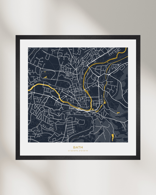 A framed minimalist street map print of Bath, England, featuring a dark navy blue background with white road lines and gold highlights, set in a black frame with a white mat against a light gray wall with soft shadows.