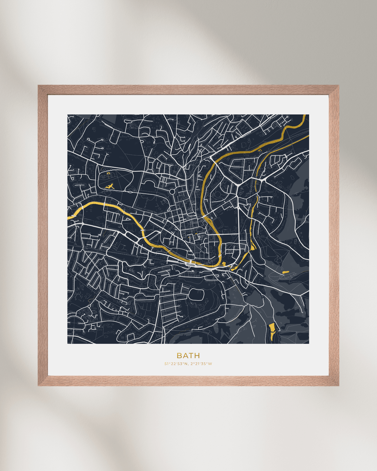 A framed street map art print of Bath, UK, featuring a dark navy blue background with white street lines and waterways highlighted in gold foil. The print is matted in white and housed in a light natural wood frame, shown against a neutral beige wall with soft diagonal shadows.