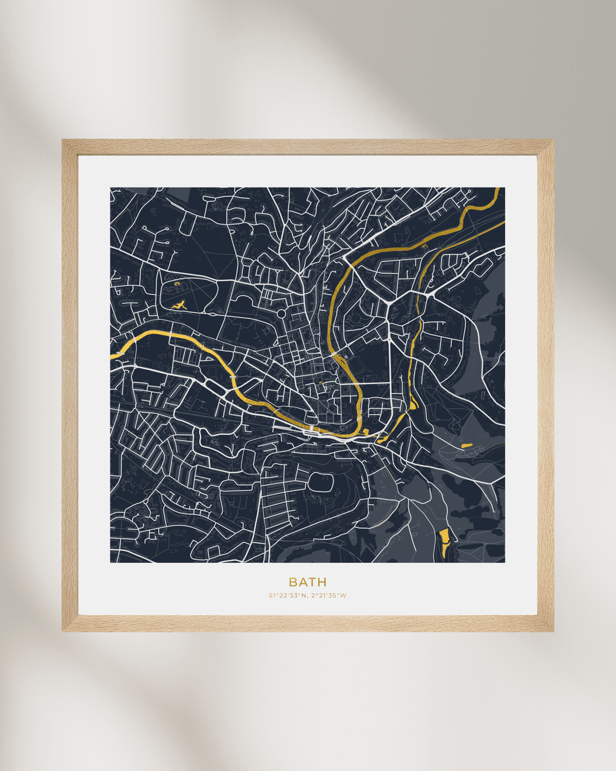 A framed city map print of Bath, UK, featuring a dark navy blue background with white street lines and waterways highlighted in gold foil. The print is styled with a wide white mat and a light natural wood frame, set against a neutral light gray wall with soft leaf shadows.