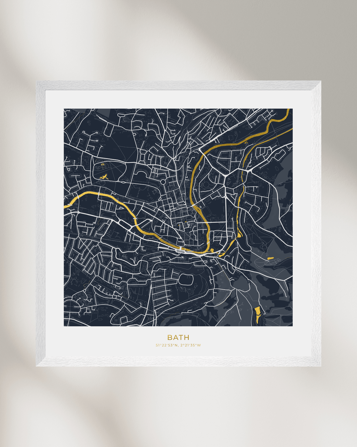 A framed city map print of Bath, UK, featuring a dark navy blue background with white street lines and waterways highlighted in gold foil. The print is styled with a wide white mat and a light natural wood frame, set against a neutral light gray wall with soft leaf shadows.