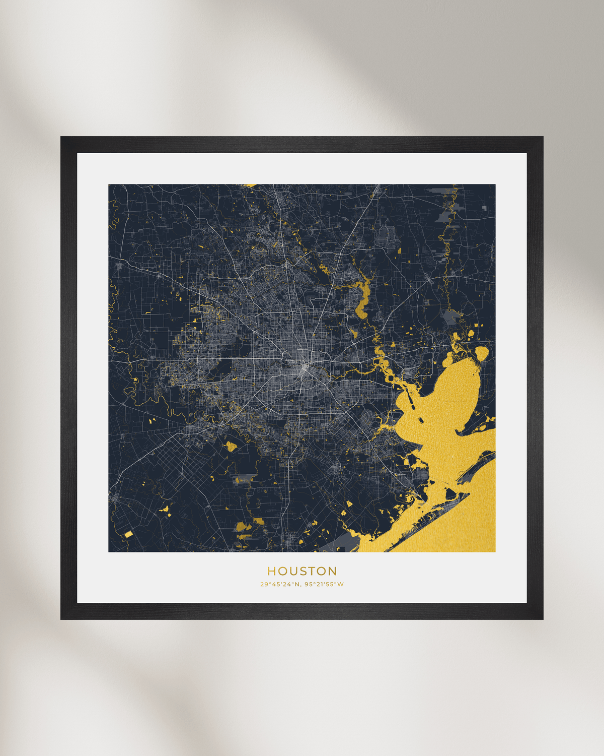 A framed Houston city map art print with a dark navy background and gold highlighted waterways, a white mat, and a thin black frame, shown against a light neutral wall with soft shadows.