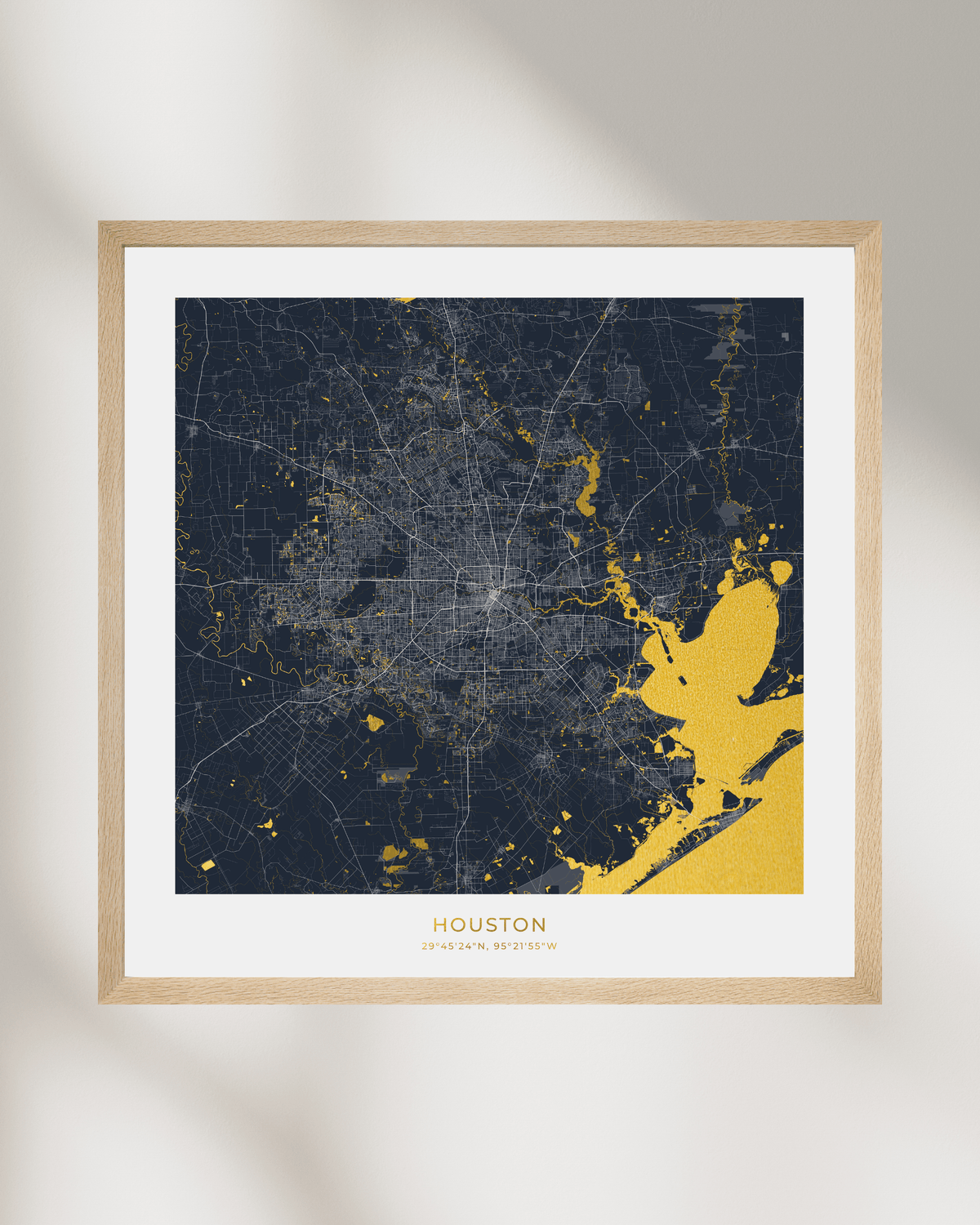 A framed Houston city map art print with a dark navy background and gold highlighted waterways, a white mat, and a thin natural wood frame, shown against a light neutral wall with soft shadows.