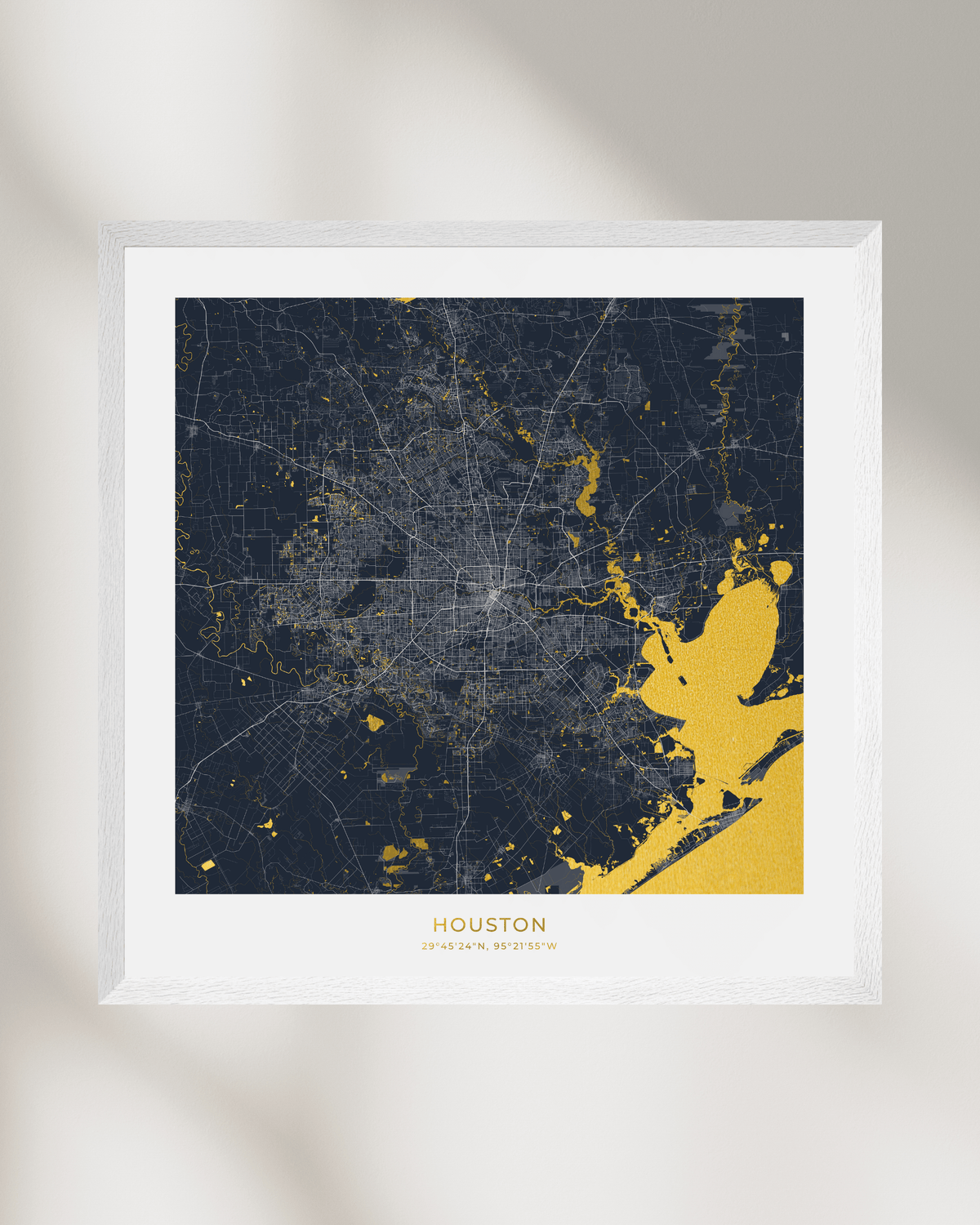 A framed Houston city map art print with a dark navy background and gold highlighted waterways, a white mat, and a thin white wood frame, shown against a light neutral wall with soft shadows.