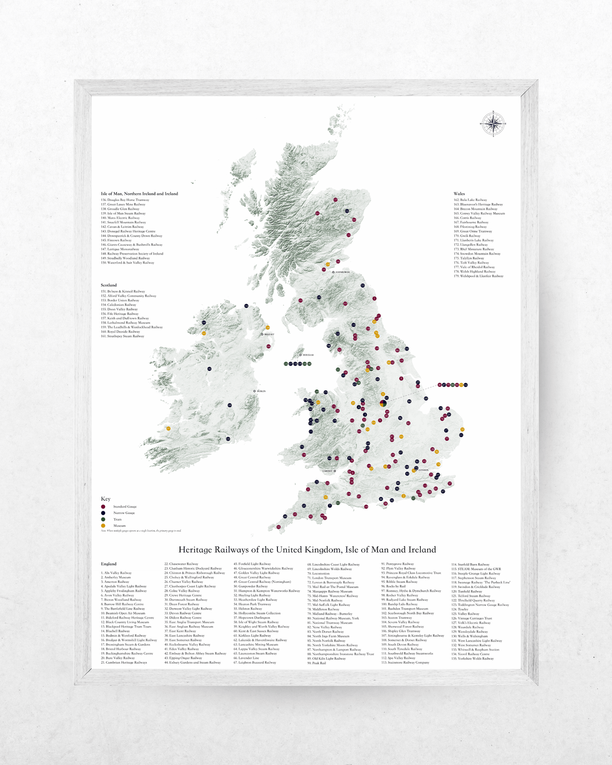 A framed poster print of a map of the United Kingdom, Ireland, and Isle of Man, featuring numerous colored dots marking heritage railway stations, with extensive station lists printed around the map. The print is housed in a light-colored wooden frame and displayed against a textured white wall.