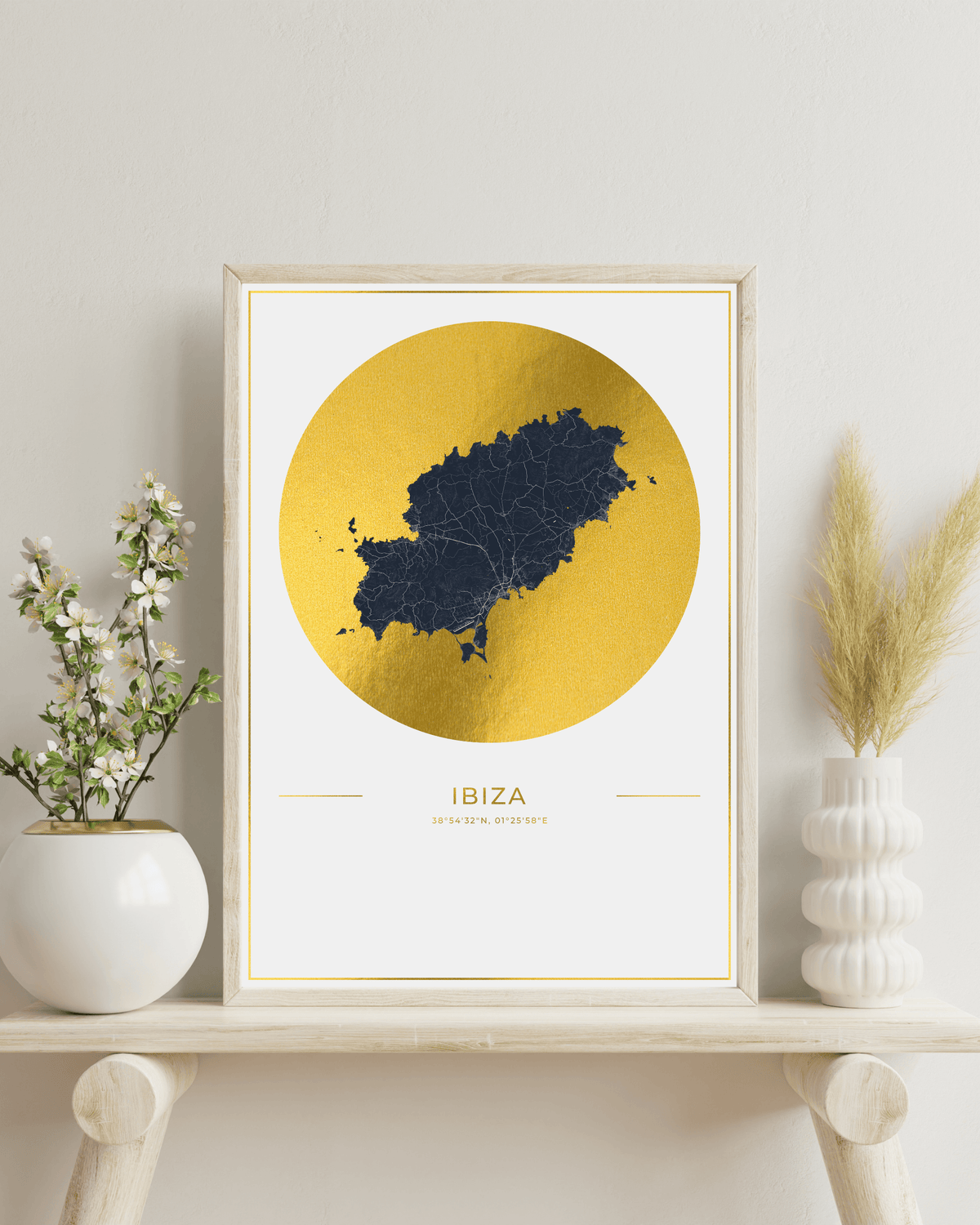A framed art print of the map of Ibiza, Spain, featuring a dark navy blue map silhouette inside a textured gold circular design on a white background. The print is housed in a light wood frame and styled on a wooden shelf next to white ceramic vases with green and beige dried plants.