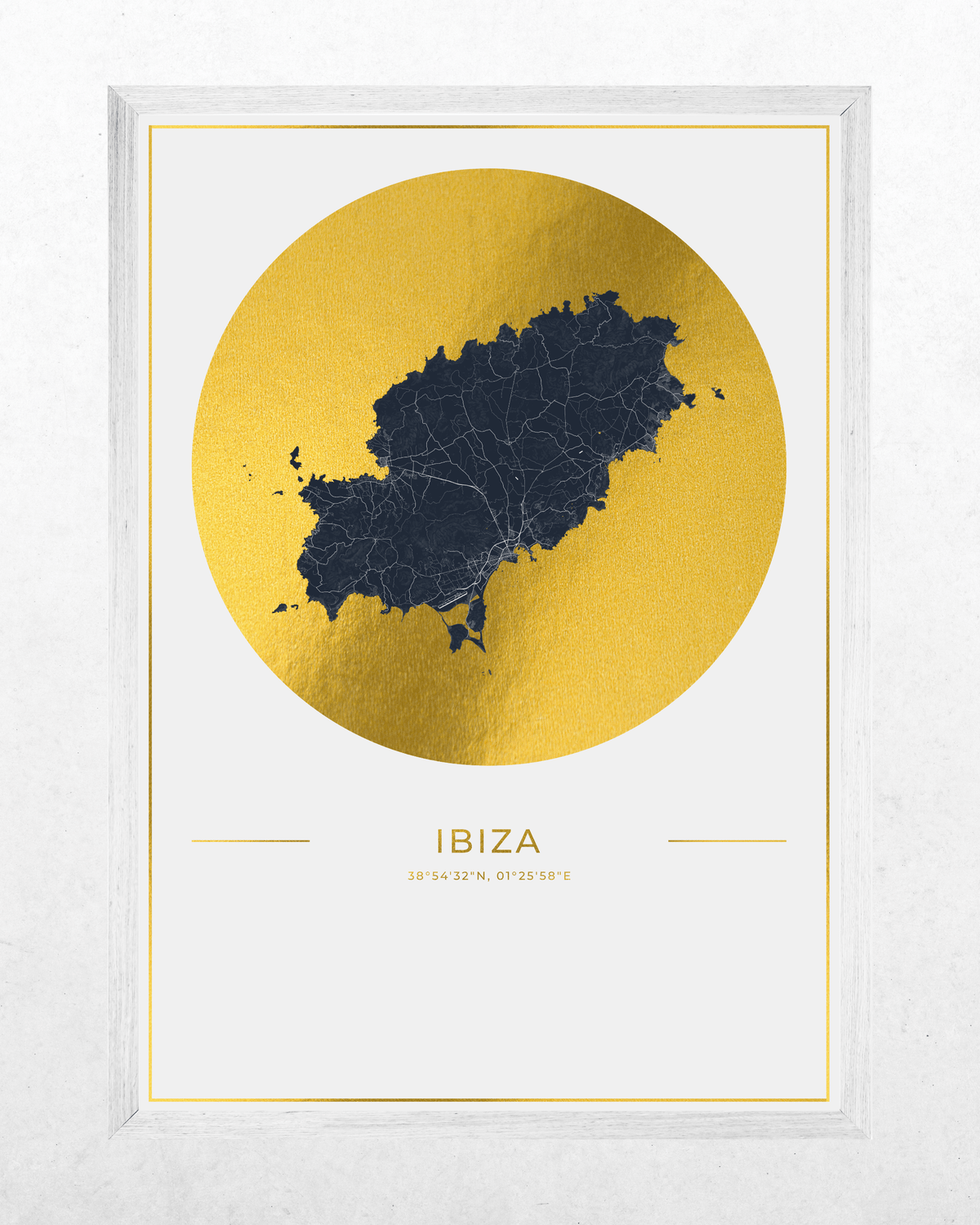 A framed minimalist art print of an Ibiza map, featuring a dark navy blue map silhouette inside a large textured gold circle on a white background, with the word 'IBIZA' and coordinates printed below. The print is housed in a white frame and set against a light gray textured wall.