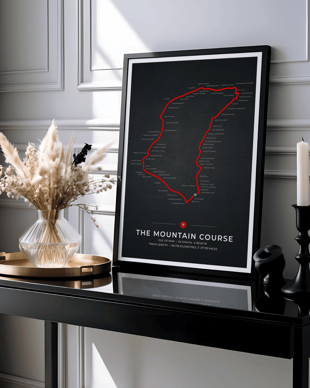 A framed poster print leaning on a black console table in a styled interior. The poster shows a black background with a red outlined mountain course and the text 'THE MOUNTAIN COURSE' near the bottom.