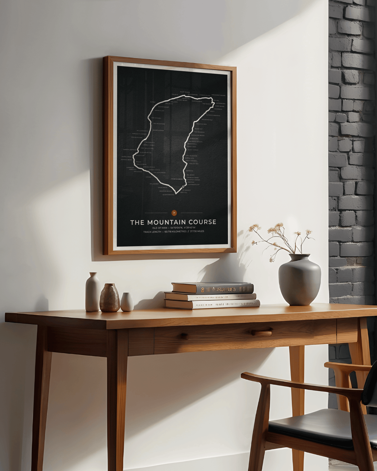 A framed poster featuring a red outline map of the Isle of Man TT race track on a black background, hanging on a white wall above a wooden desk with books and vases.
