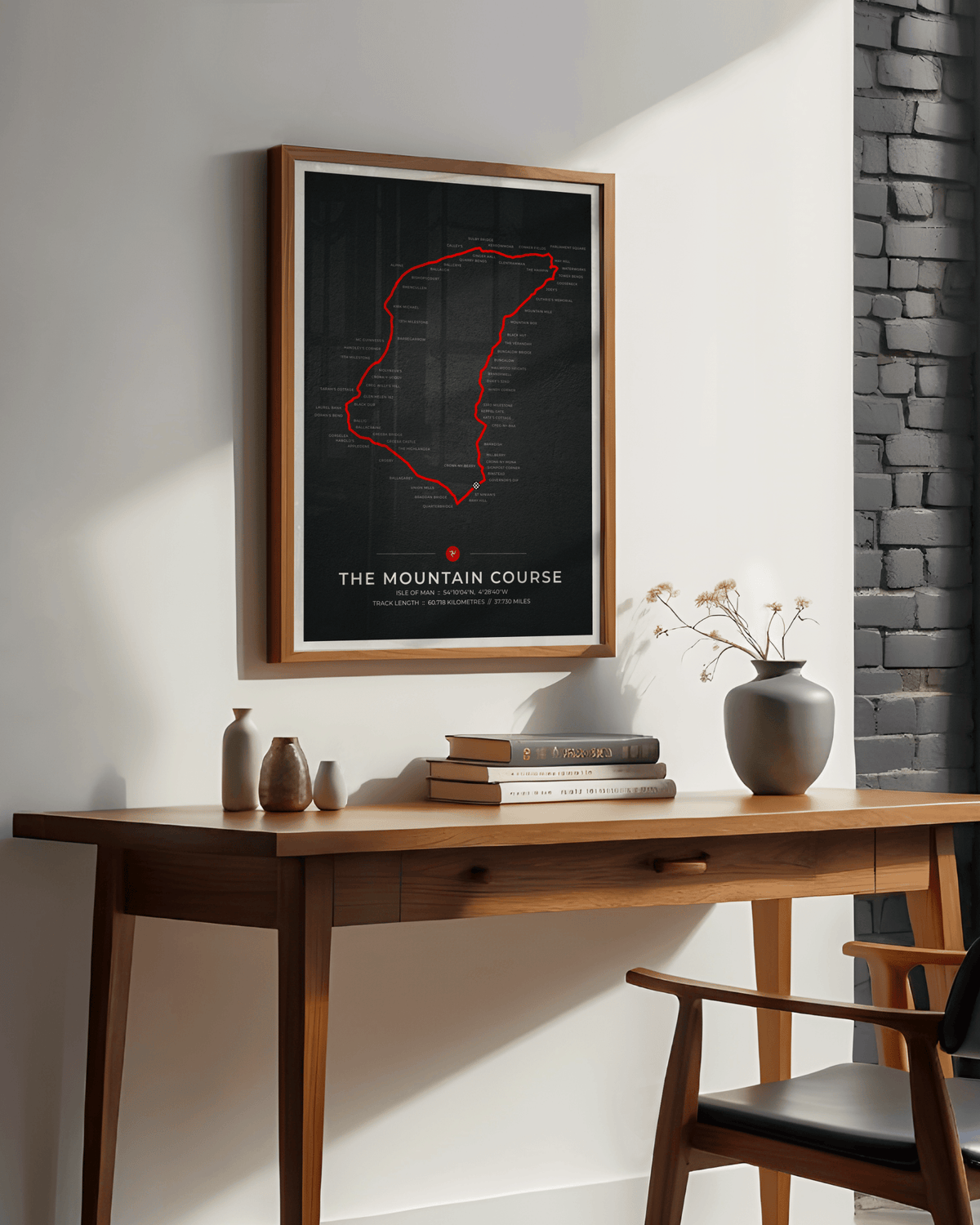 A framed poster featuring a red outline map of the Isle of Man TT race track on a black background, hanging on a white wall above a wooden desk with books and vases.