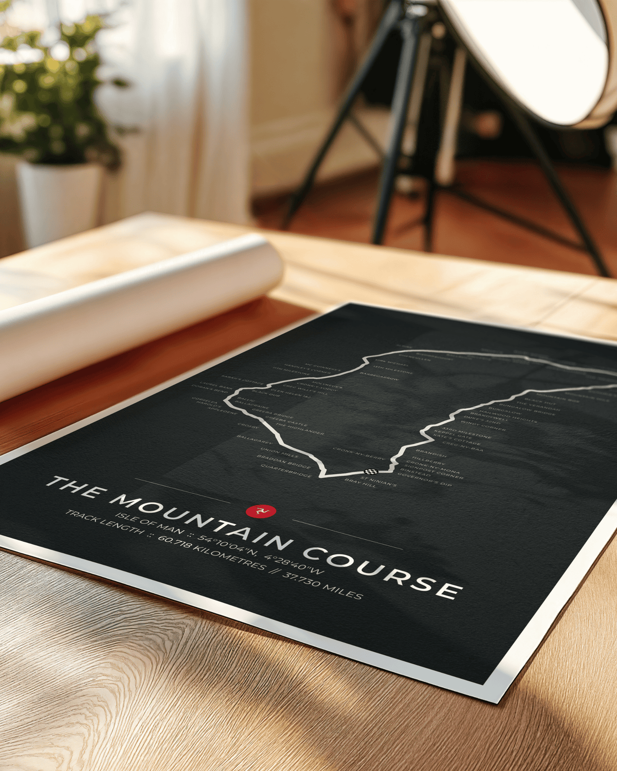 A black poster print titled "THE MOUNTAIN COURSE" with a white track outline and small red marker, placed on a wooden tabletop in a softly lit room. A rolled sheet and studio light stand are blurred in the background.