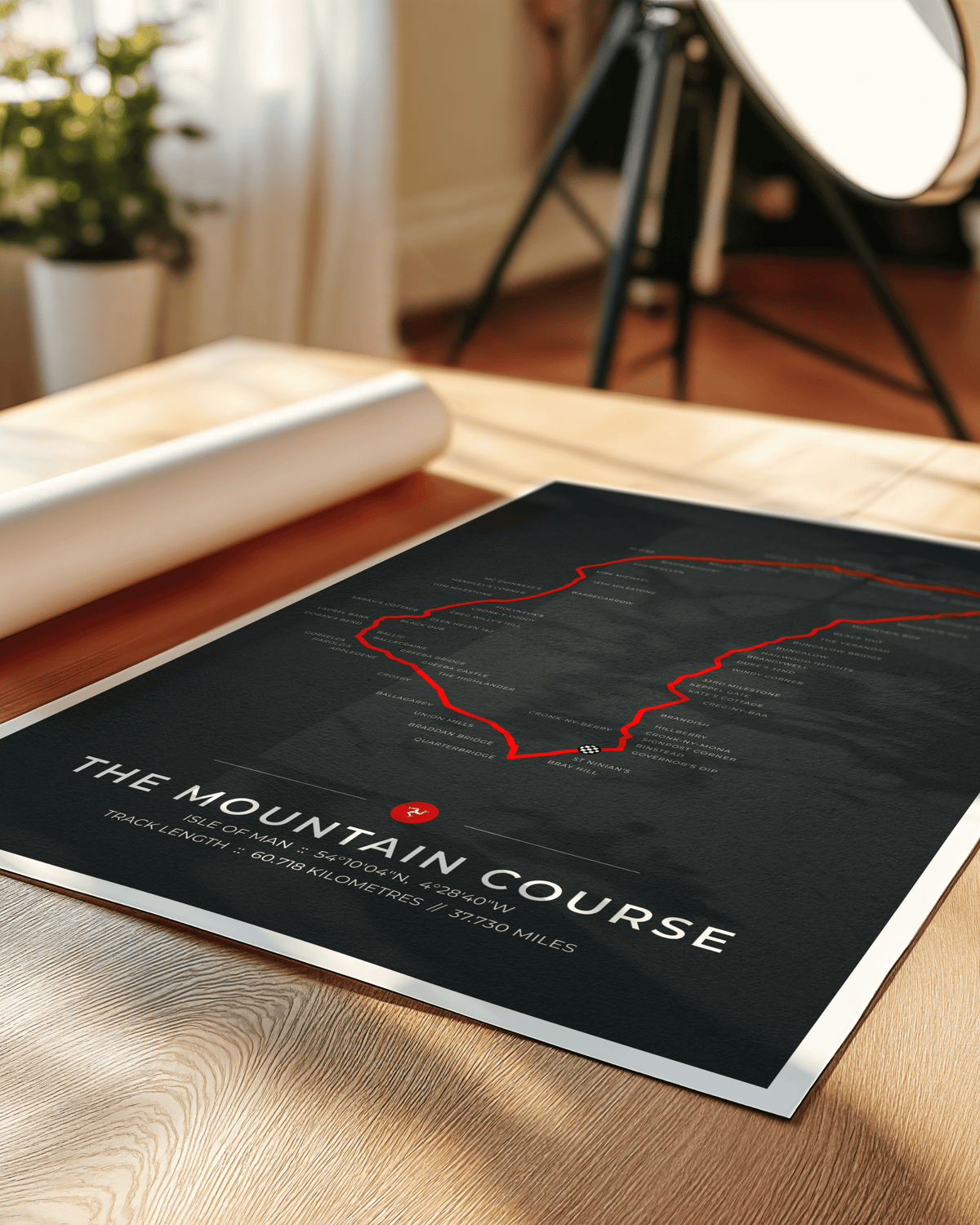 A black poster print titled "THE MOUNTAIN COURSE" with a red outlined track map and white text, displayed on a wooden table in a softly lit room with a rolled mat and studio light visible in the background.