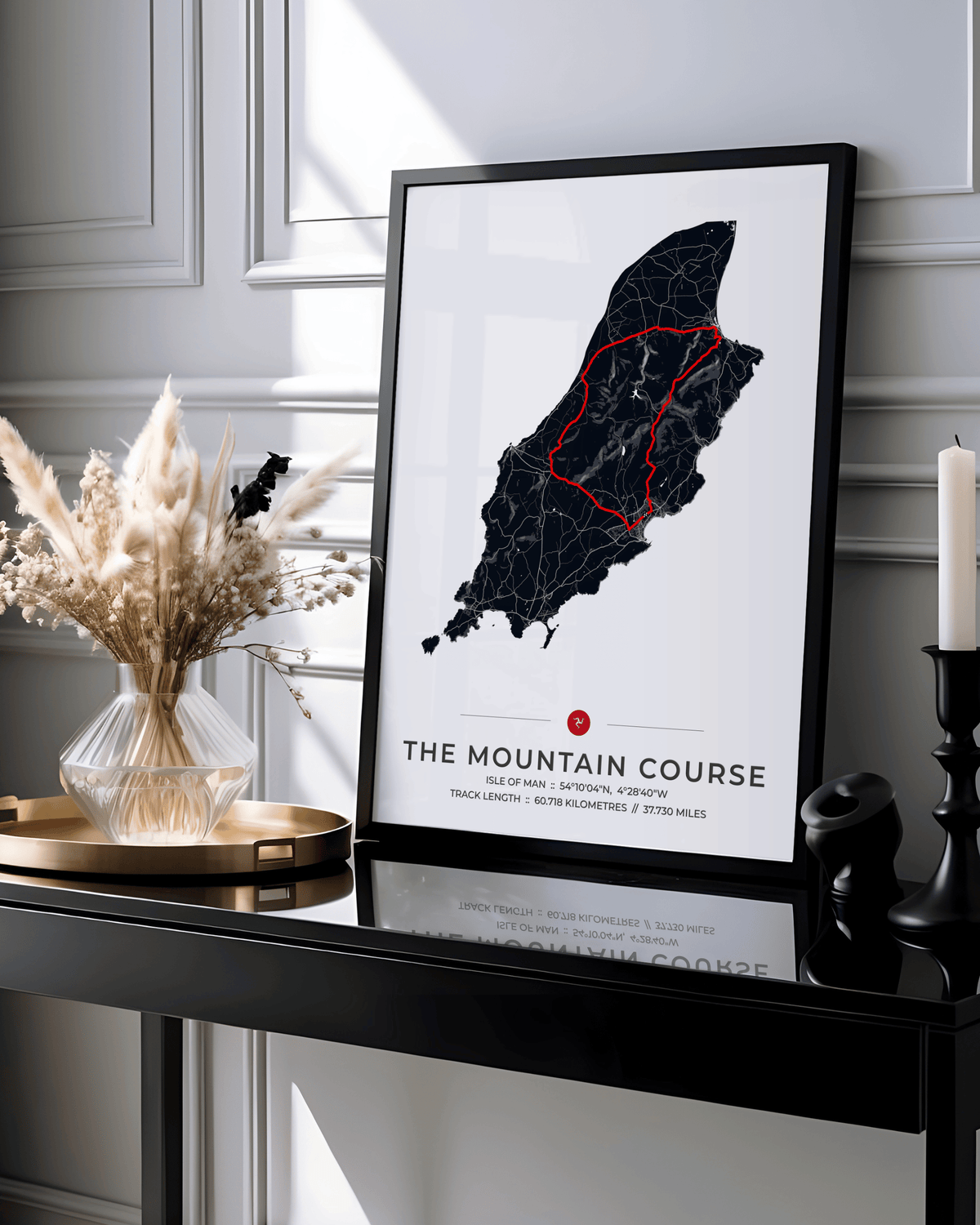 A framed minimalist art print titled 'THE MOUNTAIN COURSE' leaning on a glossy black console table. The print features a black map of a mountain range on a white background with a red highlighted section and a small red dot.