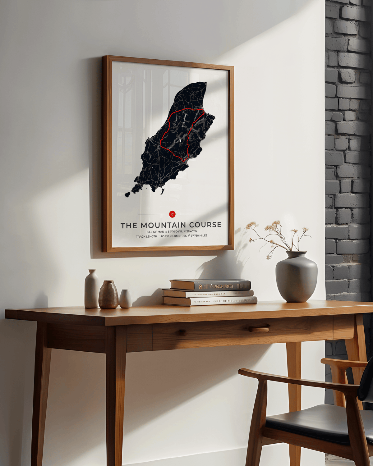 A framed art print titled 'THE MOUNTAIN COURSE' hangs on a white wall above a wooden desk with books, vases, and a chair. The print features a black map of a mountain range with a red highlighted path on a white background, enclosed in a light wood frame.