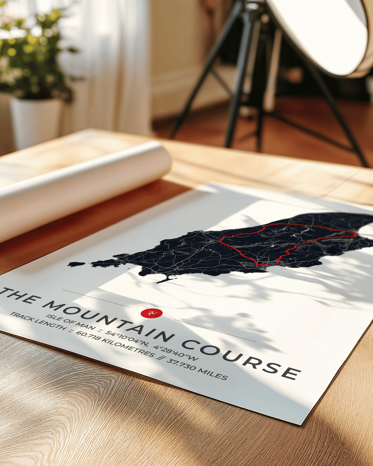 A printed poster titled "THE MOUNTAIN COURSE" lies on a wooden table in a real interior setting, with a rolled white sheet, plant, and softbox visible in the background. The poster shows a dark map of a mountain range with a red outlined track and black typography.